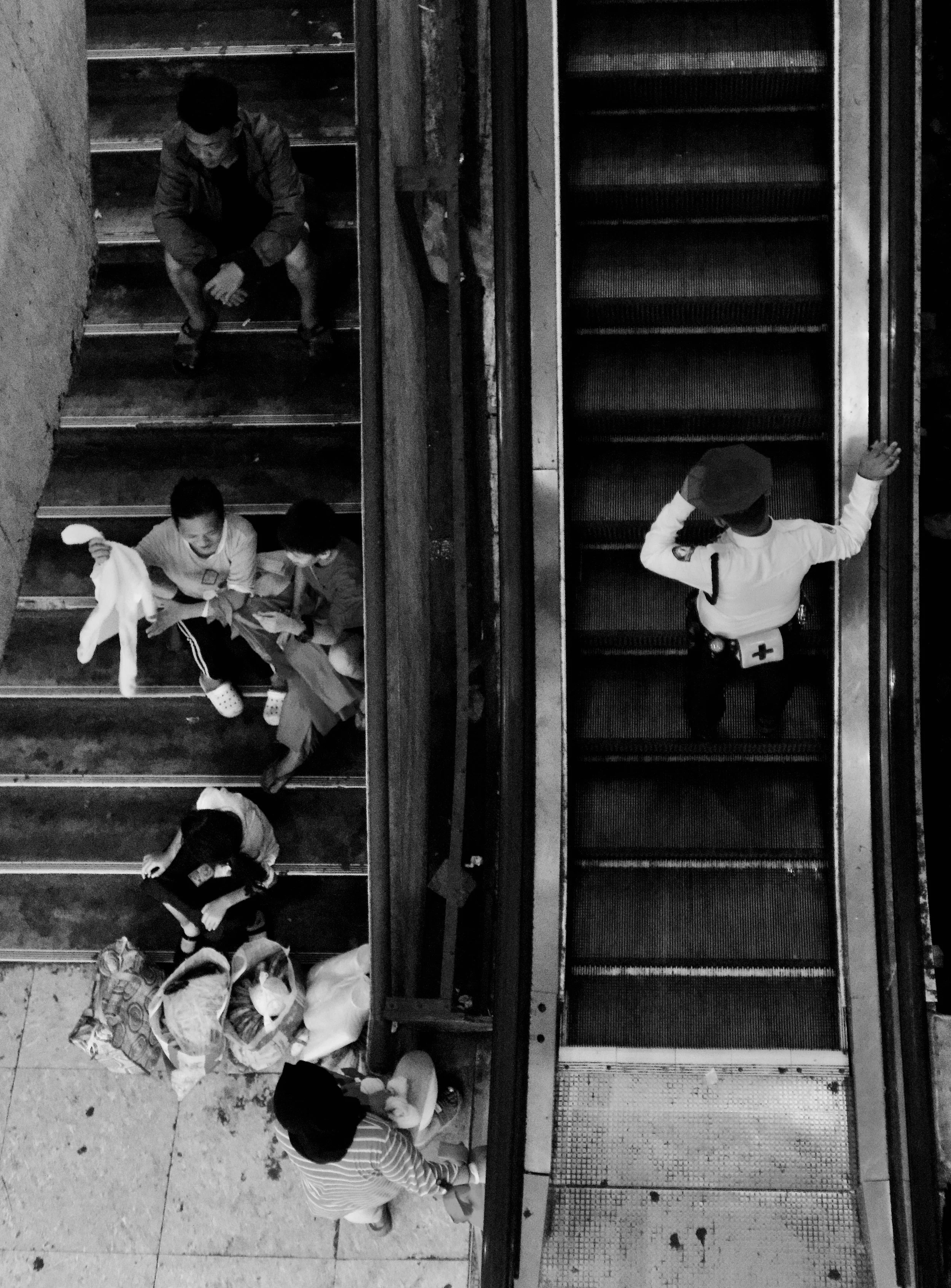 Black and white photo of people on stairs and an escalator, viewed from above. Two children are on the stairs, holding items, with a woman sitting at the bottom, talking to another woman. On the right side, a security guard is standing on the escalat