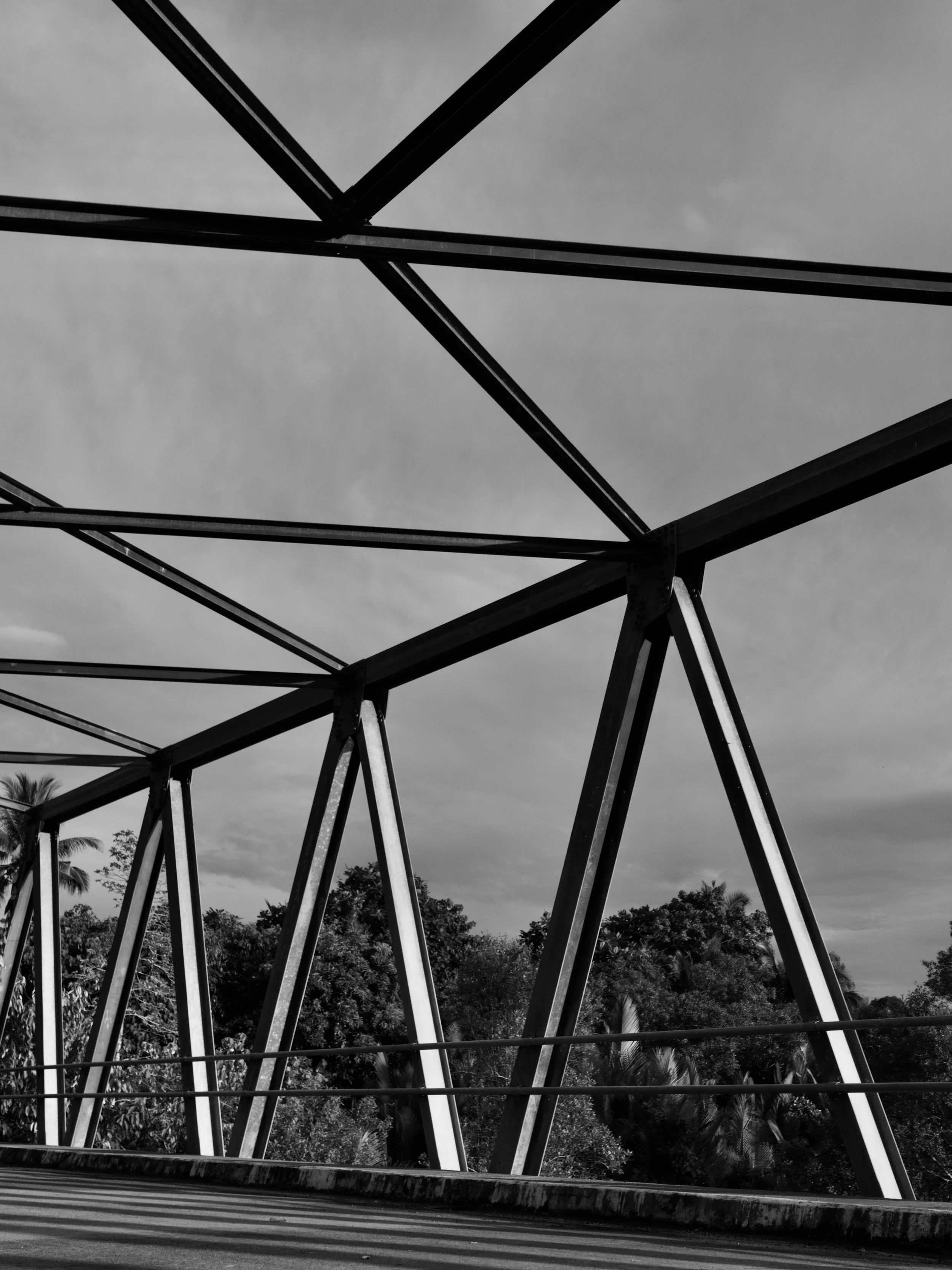 Black and white photo of a metal bridge structure with triangular supports and a cloudy sky in the background.