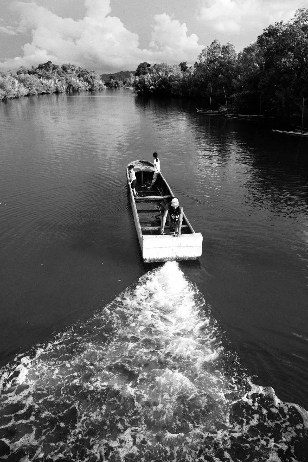 A black and white photo of a small boat on a river with three people onboard, navigating through the water surrounded by trees and clouds in the sky.