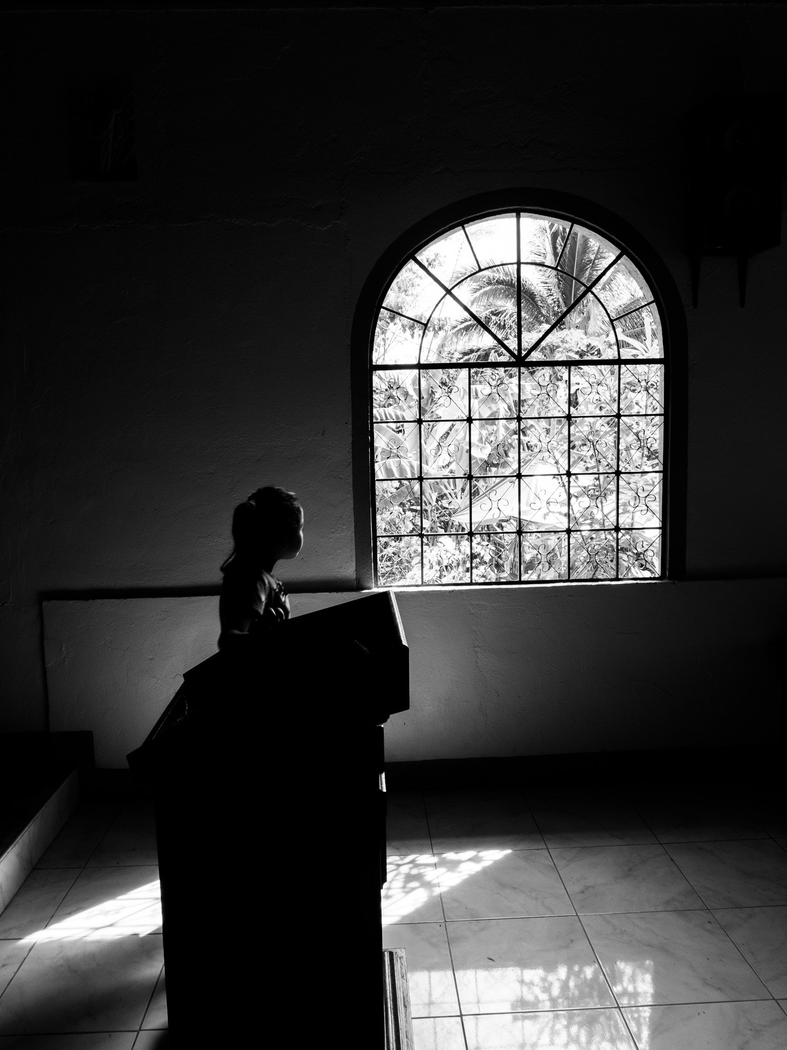 Silhouette of a person standing at a lectern near a large arched window with metal grillwork, outside trees are visible, and sunlight casts shadows on the tiled floor.
