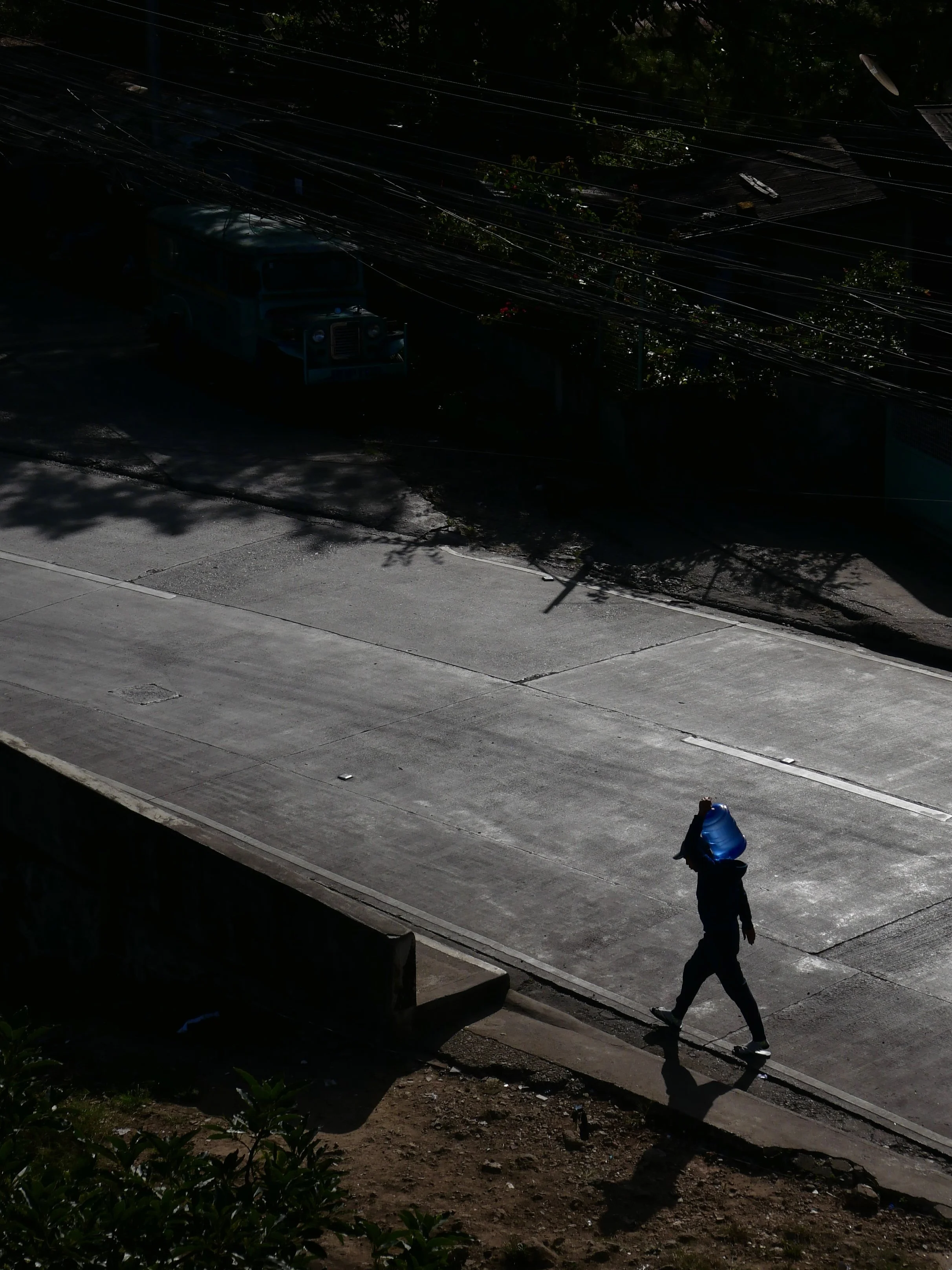 A person walking on a sunlit sidewalk carrying a blue bag, casting a shadow, with dark shaded street and overhead power lines in the background.