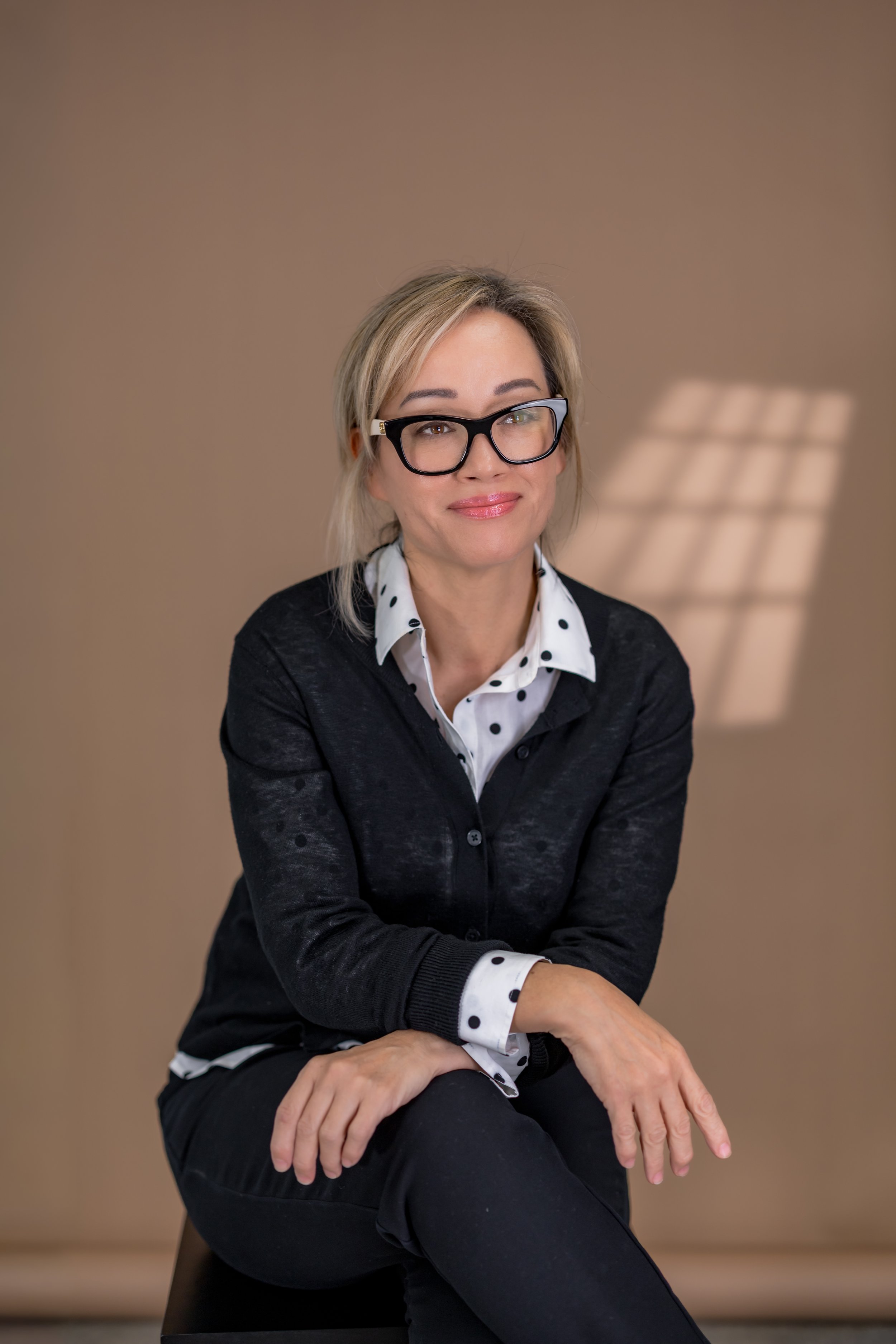 Woman wearing black cardigan and glasses seated against Beige Savage seamless backdrop with window light effect