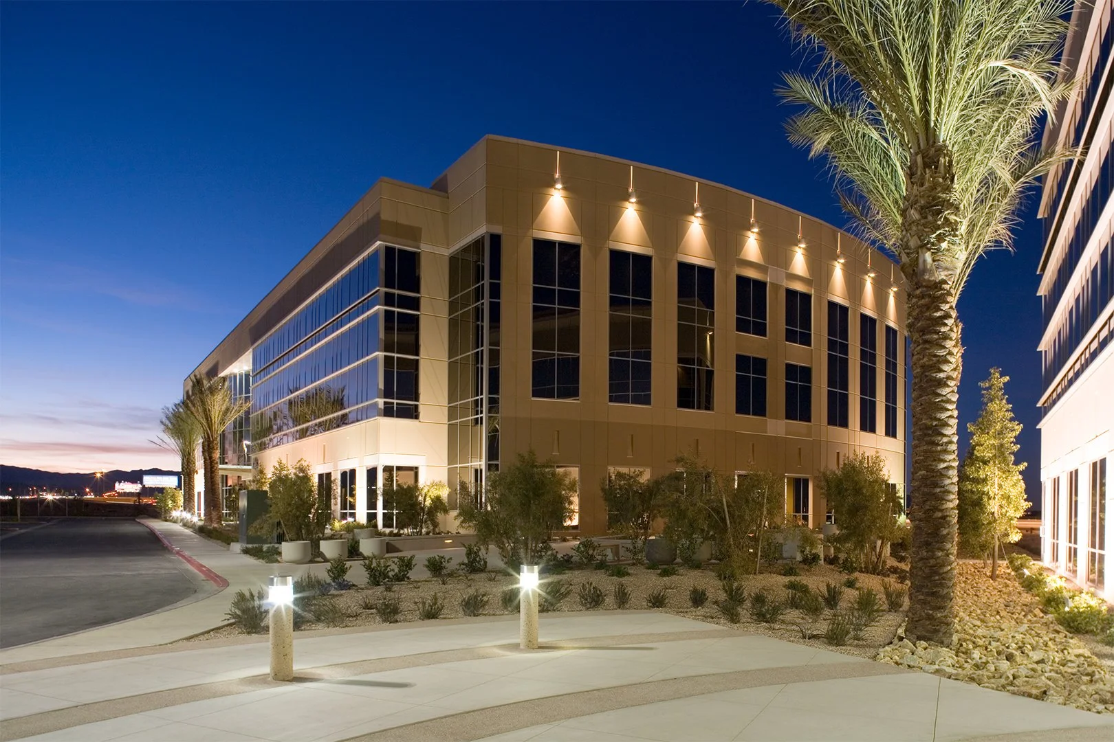 Modern office building with reflective glass windows illuminated at night, surrounded by desert landscape and palm trees.