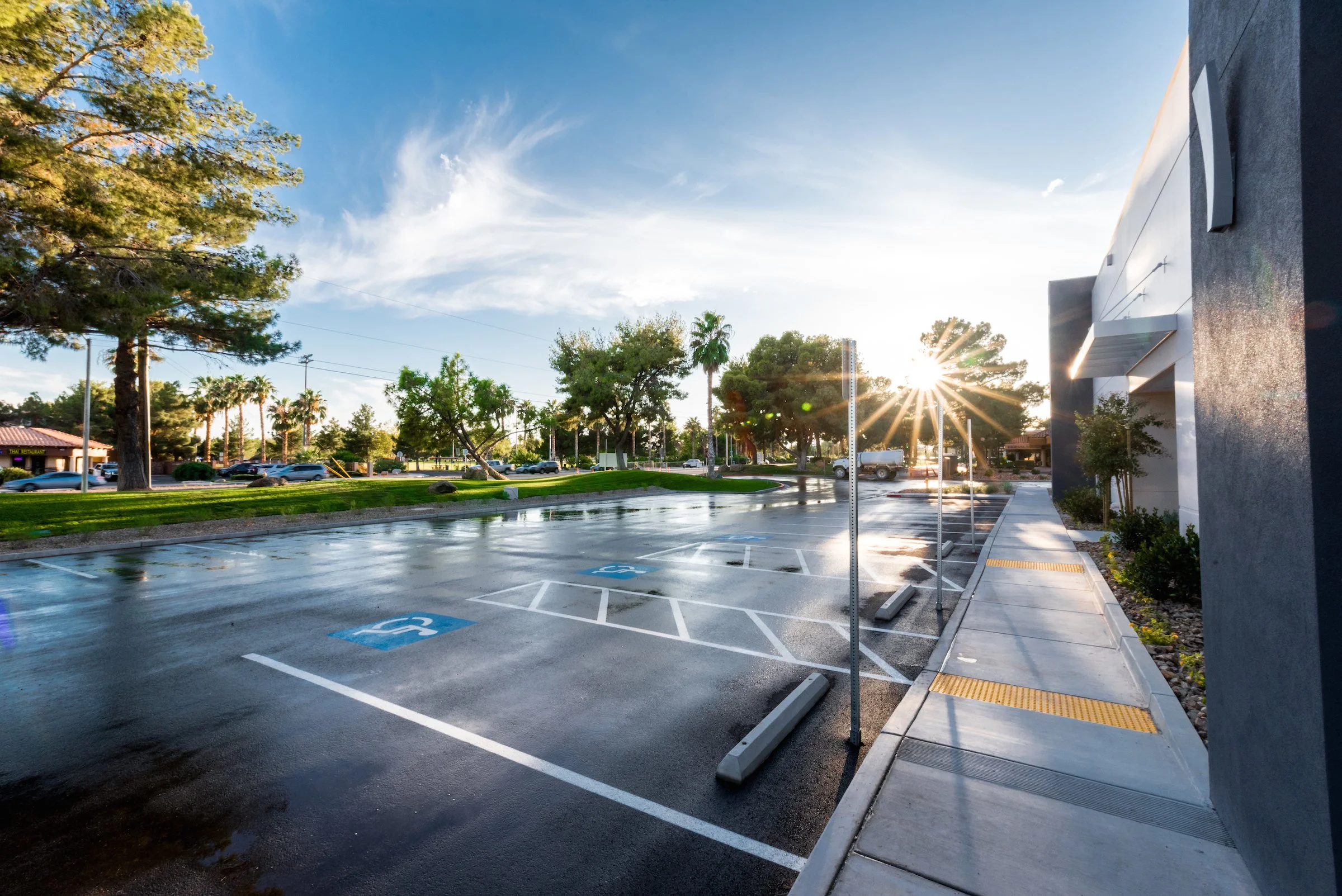 Empty parking lot with several handicapped parking spaces and a sidewalk sidewalk, sun shining through trees, wet pavement, some vehicles in distant background, with a building on the right.