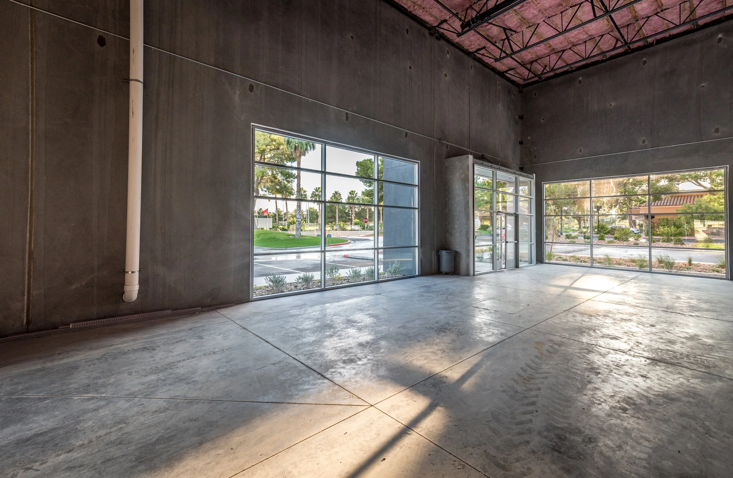 Empty commercial space or store with large windows allowing natural light, concrete floor, unfinished walls, and exposed ceiling trusses.