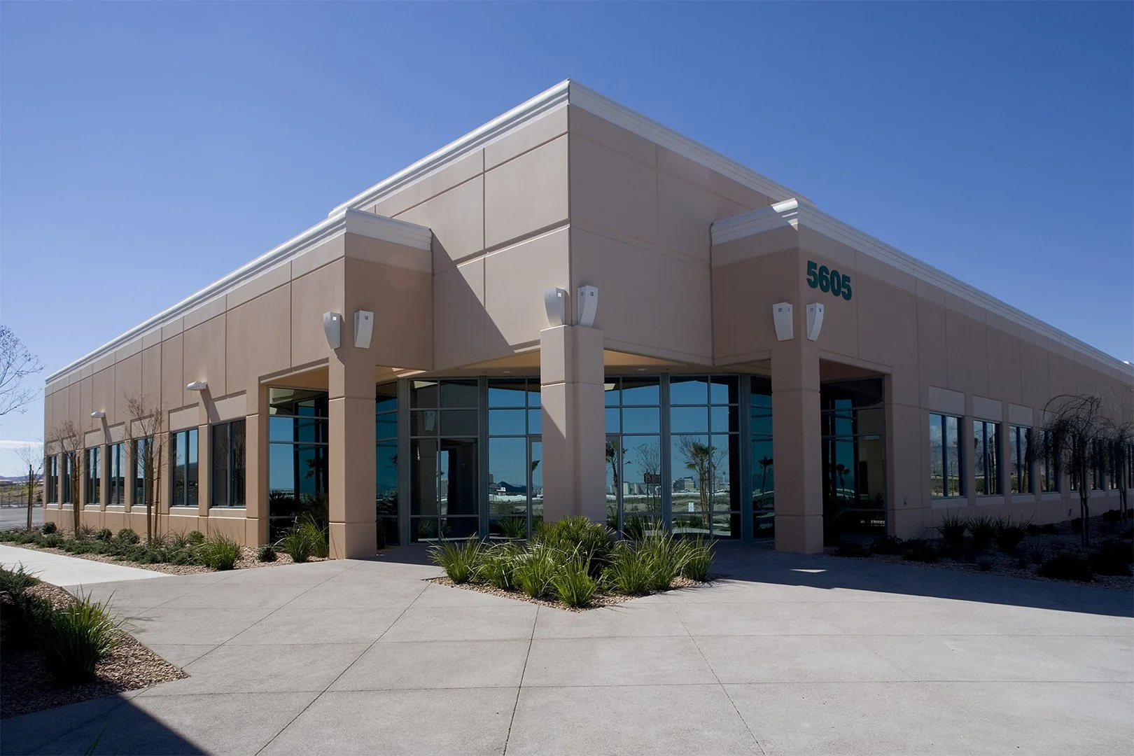 Modern commercial building with beige exterior, large windows, and the street number 5605 near the entrance, surrounded by landscaped plants and sidewalk under clear blue sky.
