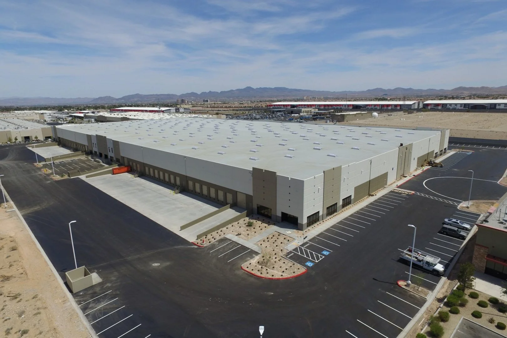 Aerial view of a large, modern industrial warehouse with parking lot and mountains in the background under a partly cloudy sky.