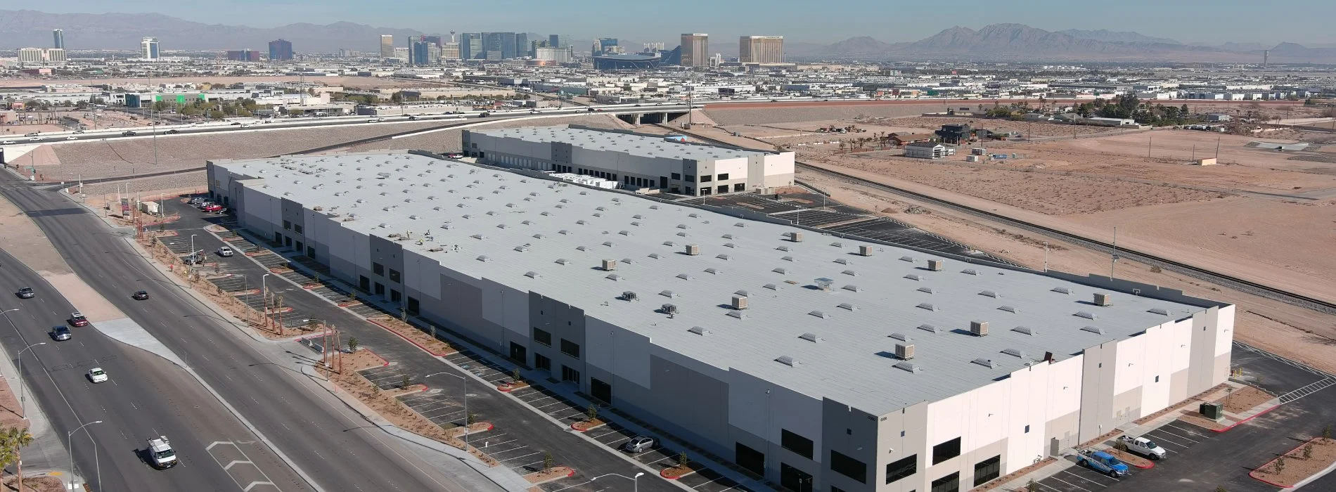 Aerial view of a large industrial building with parking lots, set in a desert area with distant mountains and a city skyline in the background.