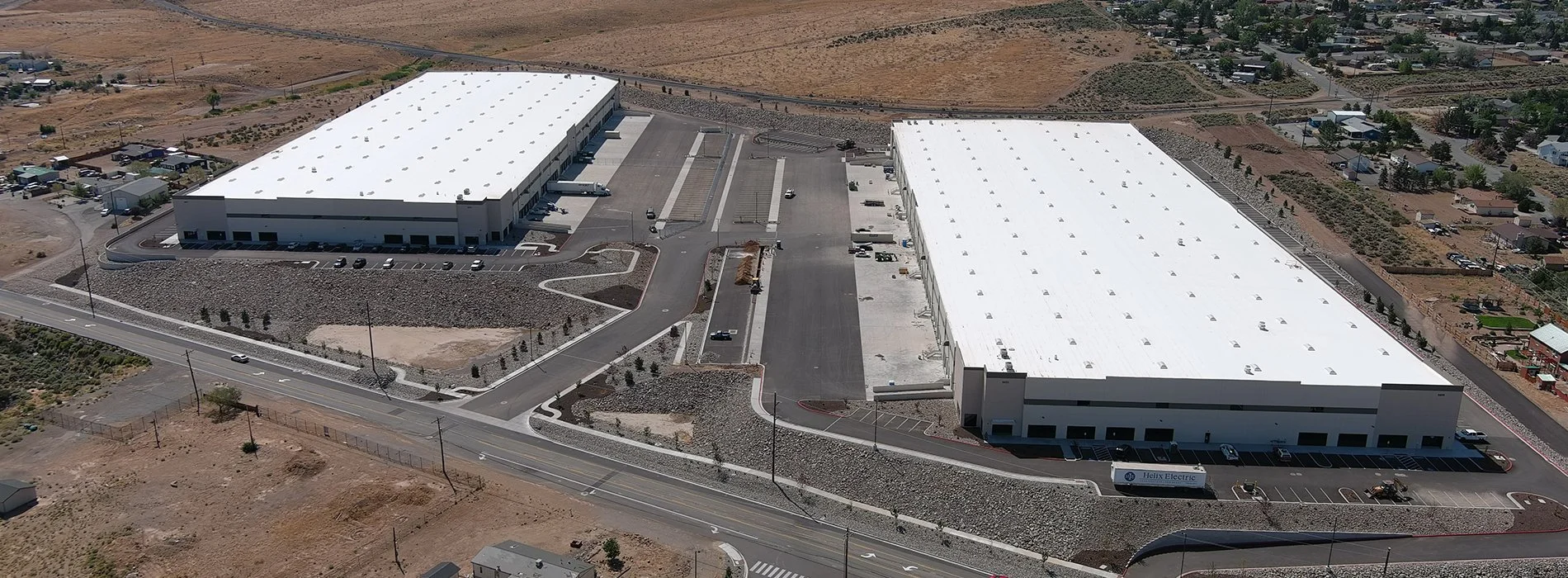Aerial view of a large industrial building with white roof, parking lot, and surrounding desert landscape.
