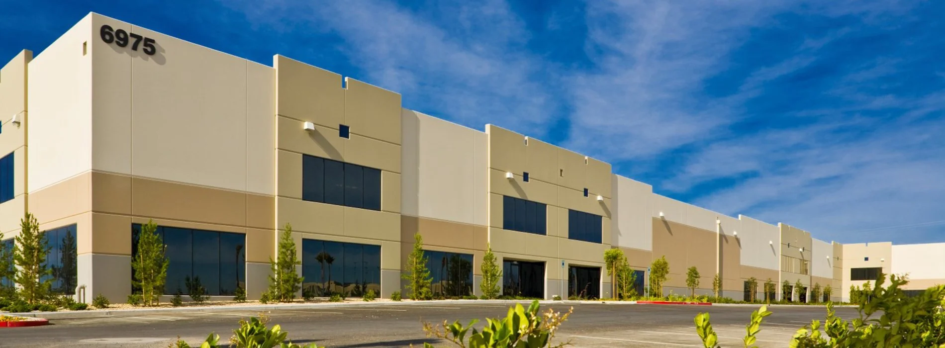 Modern commercial building with tan and white exterior, large tinted windows, greenery, and a clear blue sky.