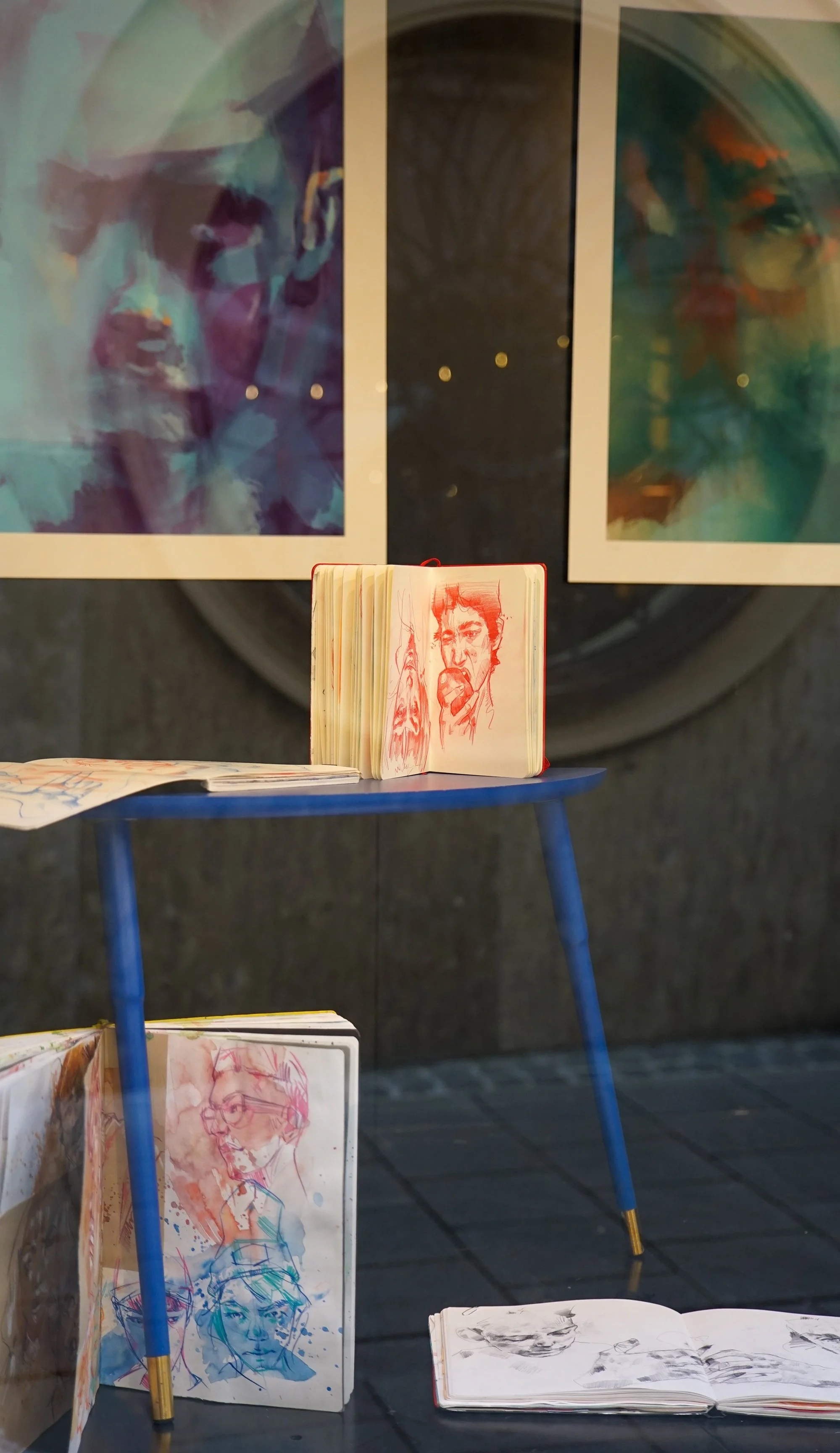 Zeichnungen und Skizzen in Skizzenbüchern, die auf einem Fenster-Display gezeigt werden, mit abstrakten und porträtartigen Kunstwerken in den Hintergründen.