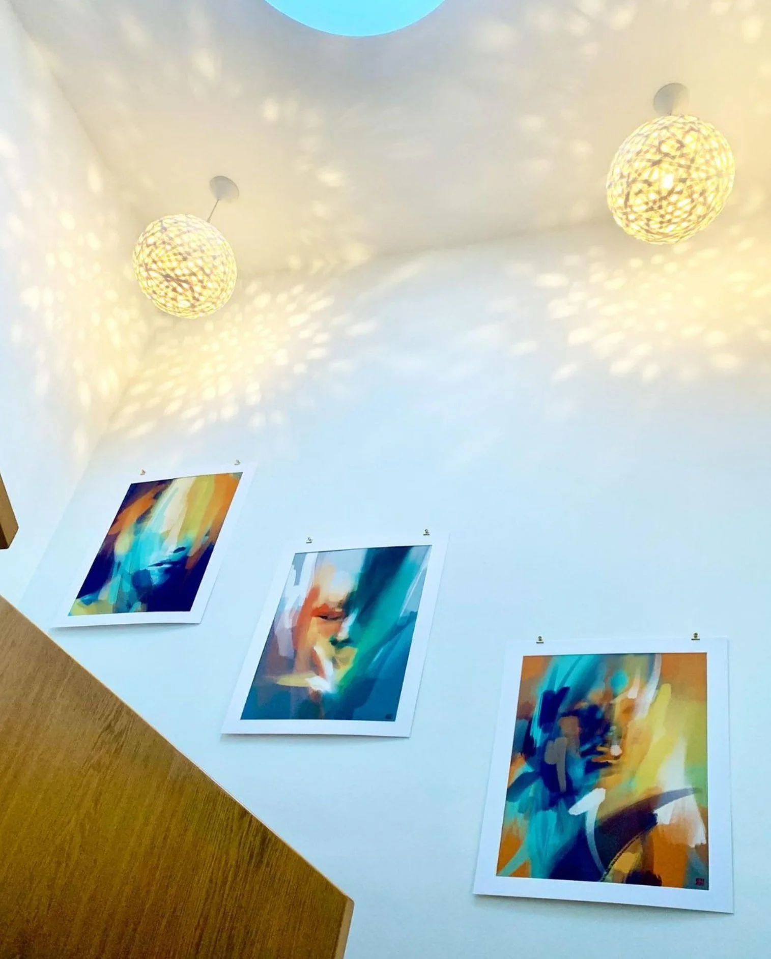 Der Raum ist hell erleuchtet und zeigt eine Decke mit zwei schwebenden, dekorativen Lampen, die Licht und Schatten an die Wände werfen. An der weißen Wand hängen bunte, abstrakte Kunstwerke, die in leuchtenden Farben gestaltet sind.