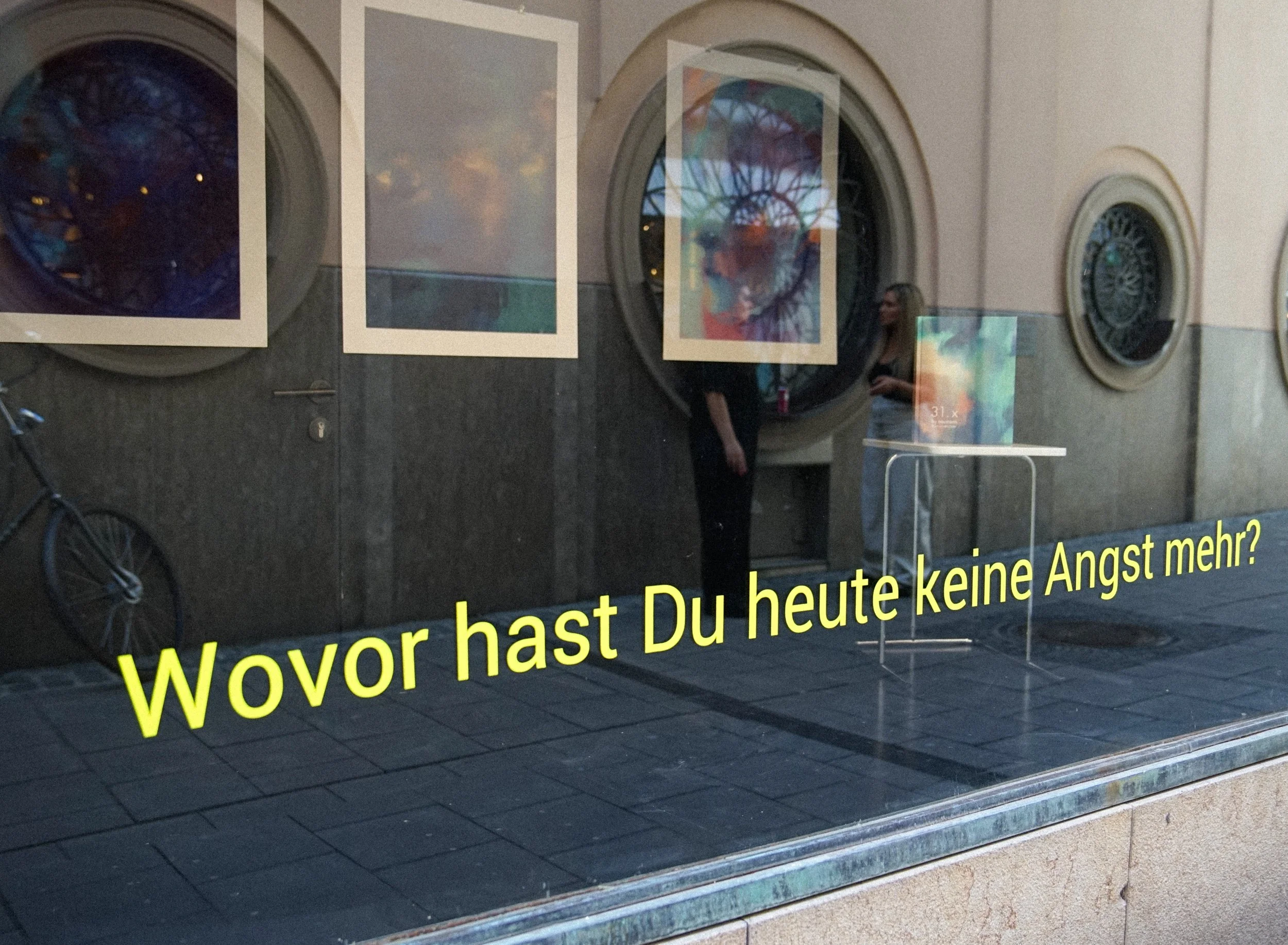 Schaufenster mit reflektierenden Bildern und gelbem Text auf dem Glas, der auf Deutsch gefragt: 'Wovor hast Du heute keine Angst mehr?'
