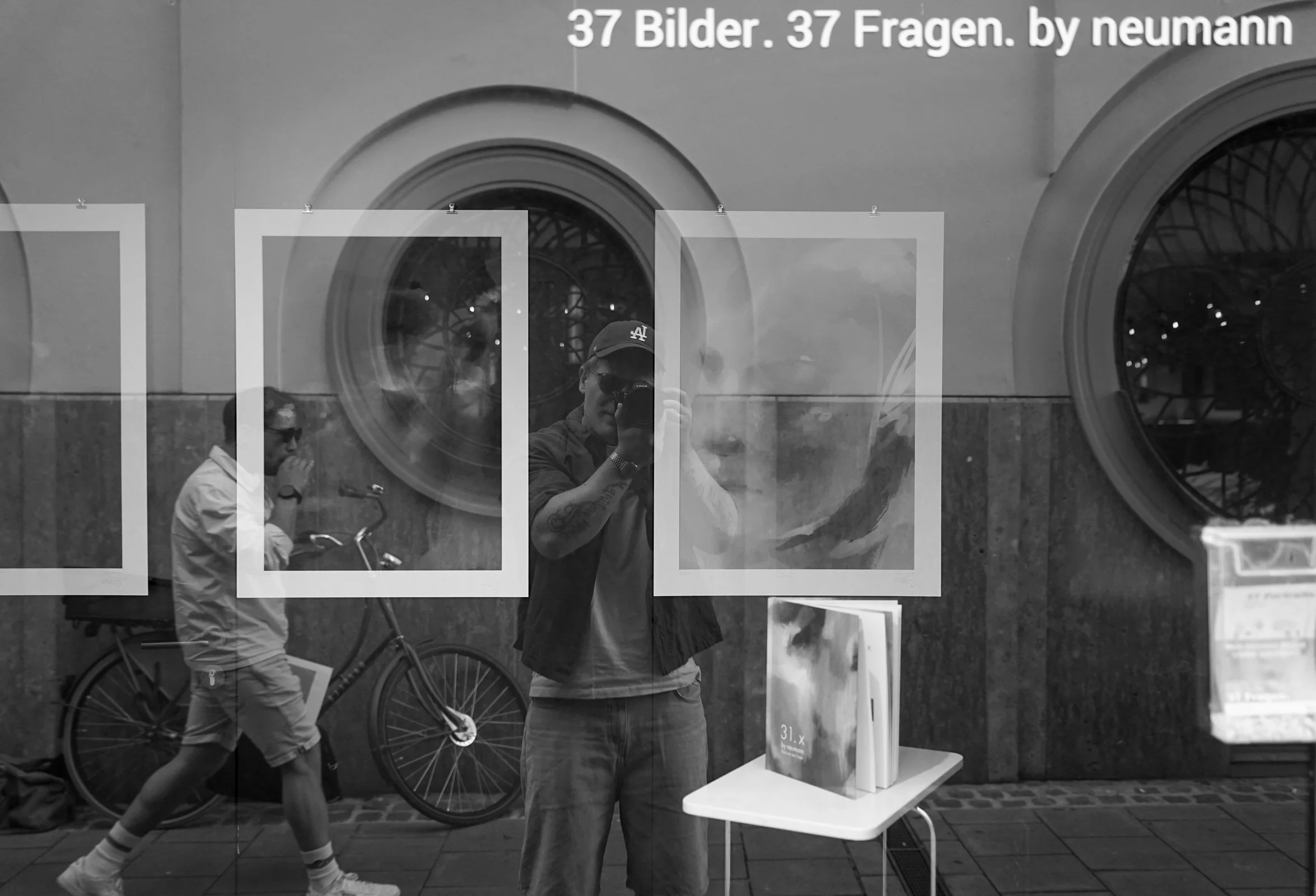 Ein Mann, der ein Foto macht, spiegelt sich in einem Fenster mit einem schwarzen Fahrrad im Hintergrund. Weitere Personen sind im Hintergrund zu sehen, sowie ein Tisch mit Büchern und eine Wand mit runden Fenstern. Es hat deutschen Text auf dem Fenster, der lautet: '37 Bilder. 37 Fragen. by neumann'.