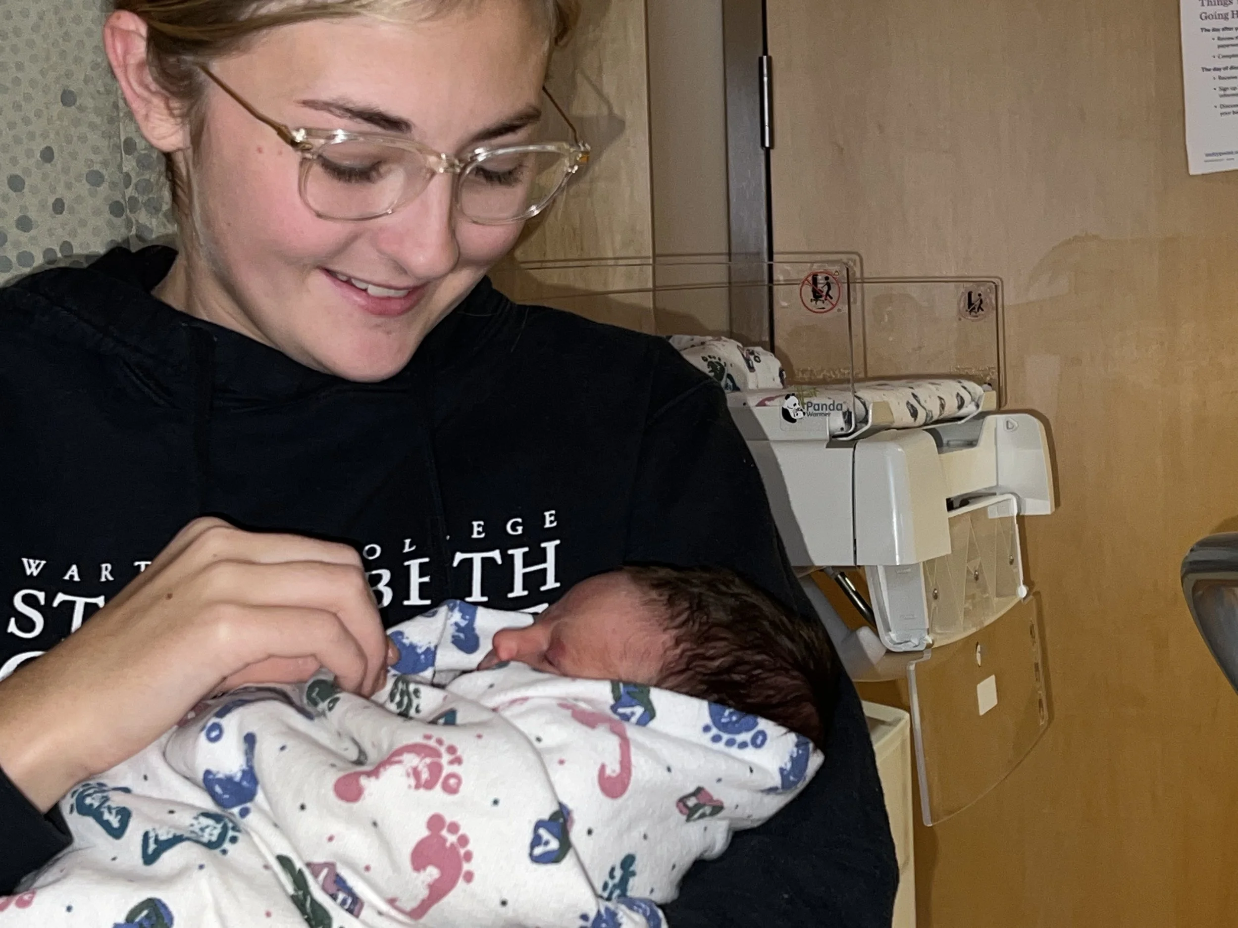 A woman wearing glasses smiling as she holds a newborn baby wrapped in a colorful blanket inside a hospital room with medical equipment and signs in the background.