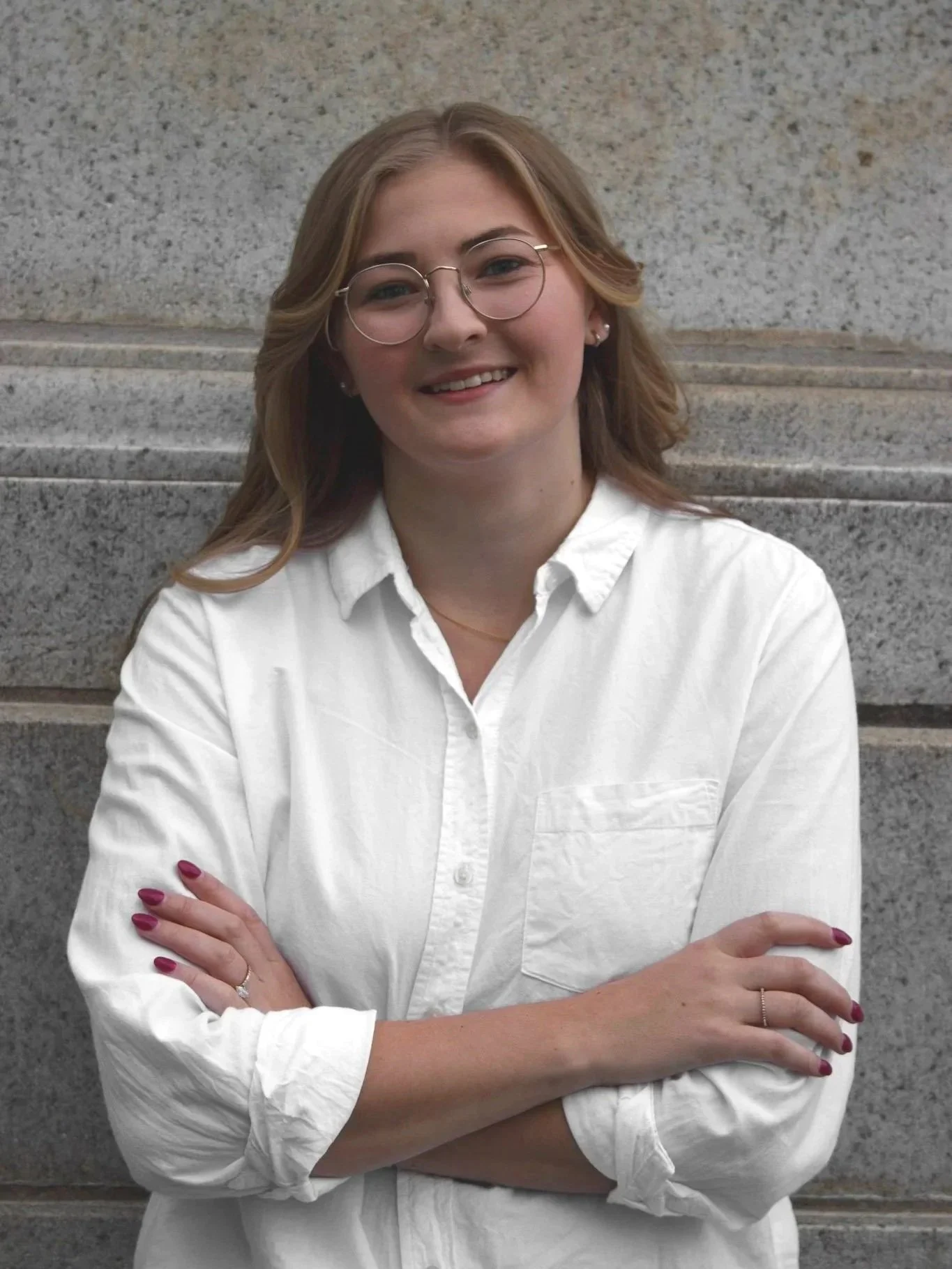 A woman with wavy blonde hair and glasses, wearing a white button-up shirt, smiling, standing in front of a stone wall.