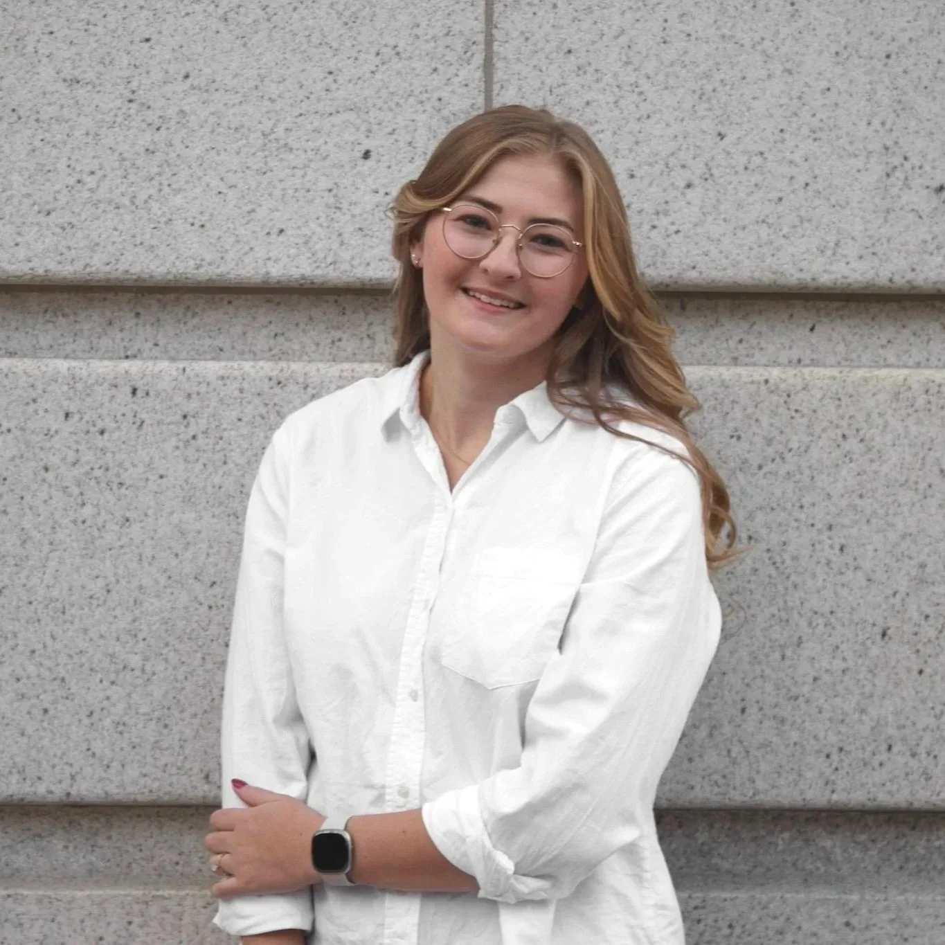 Young woman with glasses and long wavy hair wearing a white button-up shirt, standing against a gray stone wall, smiling.