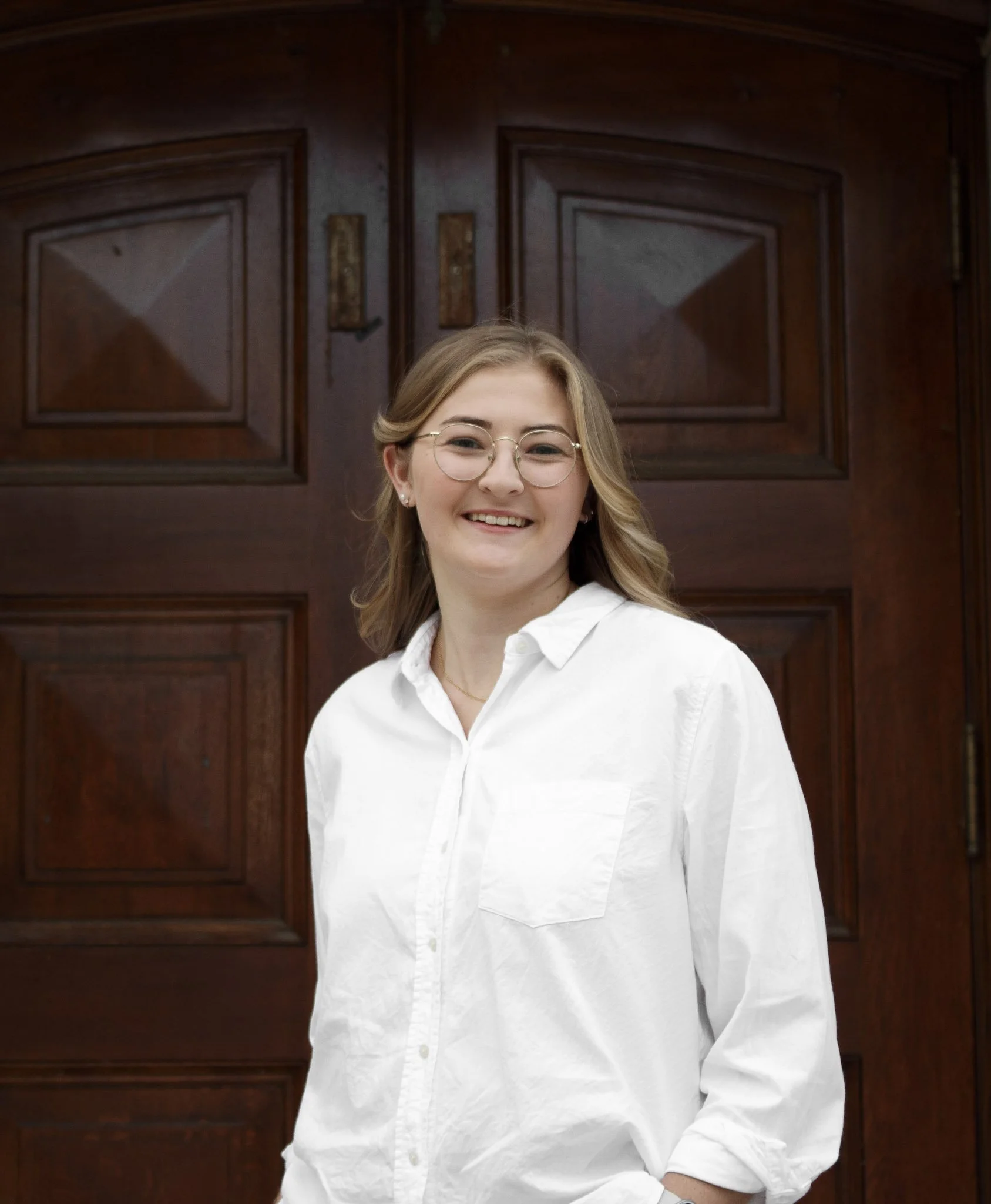 A young woman with blonde hair, eyeglasses, and earrings wearing a white button-up shirt standing in front of a dark wooden door with decorative panels.