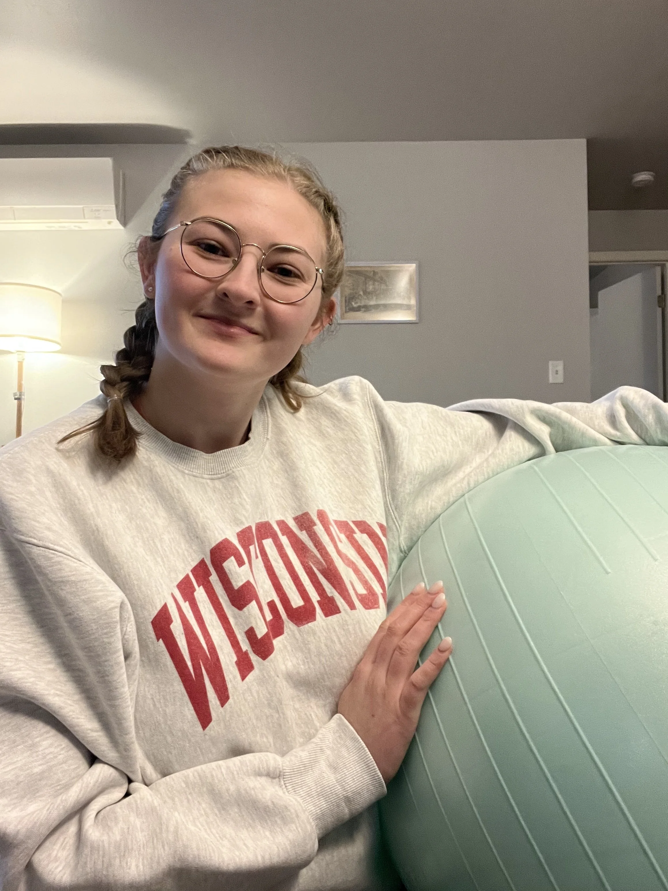 A young woman with glasses, wearing a Wisconsin sweatshirt, smiling and resting her hand on a light green exercise ball in a living room with a lamp and picture frame in the background.