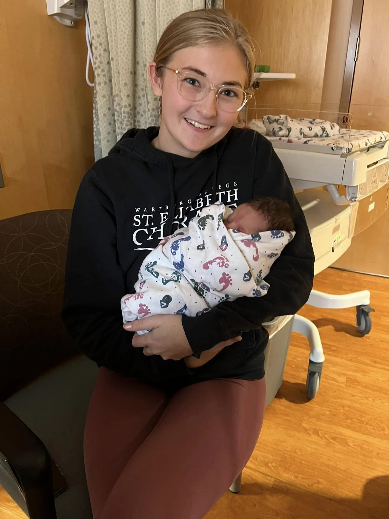 A woman sitting in a hospital room holding a newborn baby wrapped in a blanket featuring colorful footprints, with a hospital bassinet and medical equipment in the background.