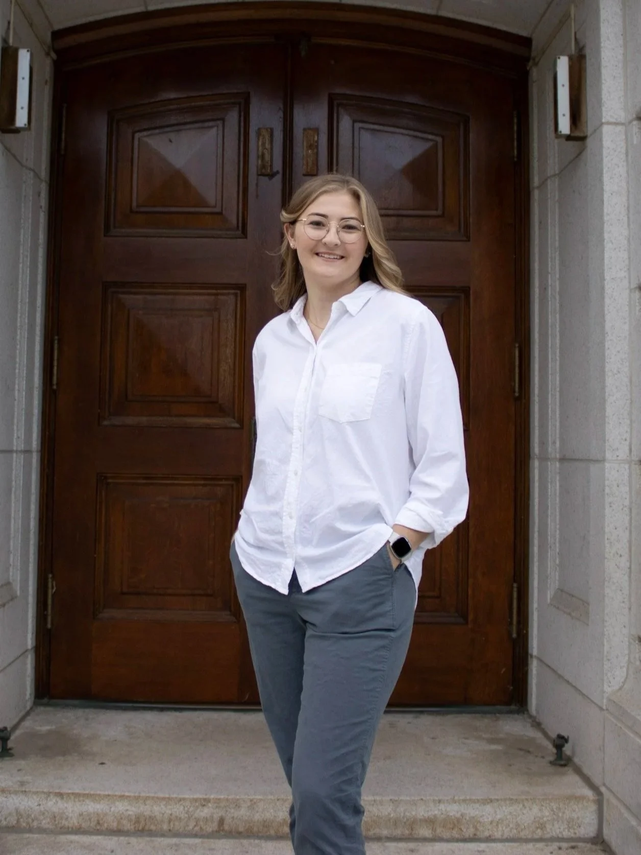 A woman with blond hair, glasses, and a watch standing in front of a large wooden door, smiling at the camera. She is wearing a white shirt and gray pants with her hands in her pockets.