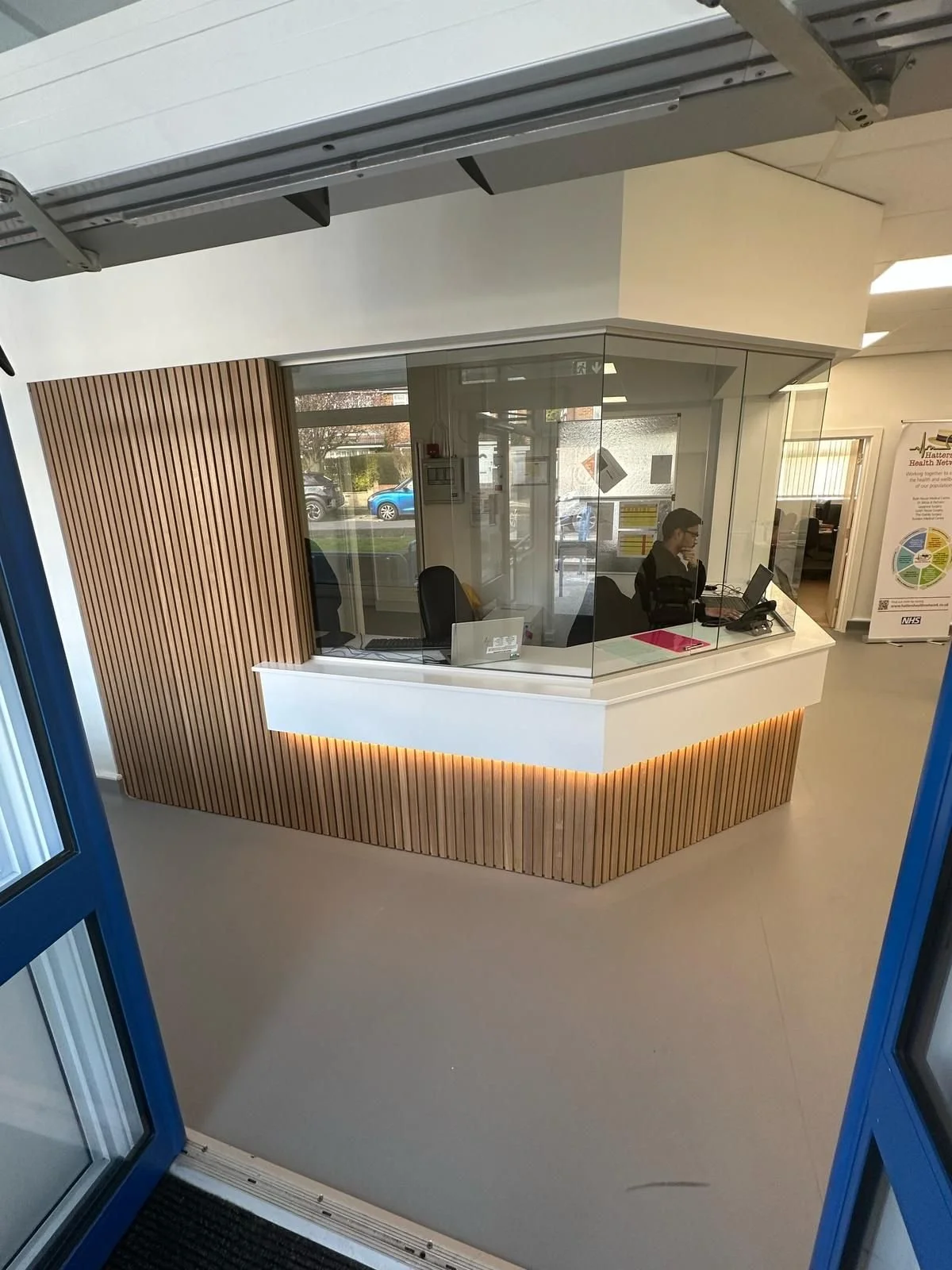 Reception area with a person at a desk, glass partition, wood paneling, and office equipment.