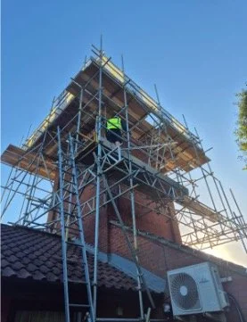 Scaffolding set up around a brick chimney on a building, with a construction worker in a yellow vest working at the top. The building has a tile roof and an air conditioning unit mounted on the wall.