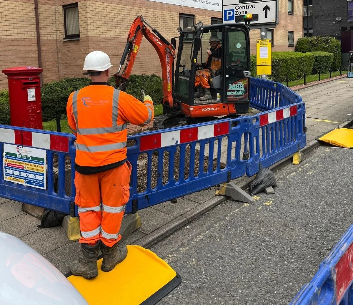 Construction workers in orange safety gear at a job site, operating a small excavator and surrounded by blue safety barriers, near a red post box.
