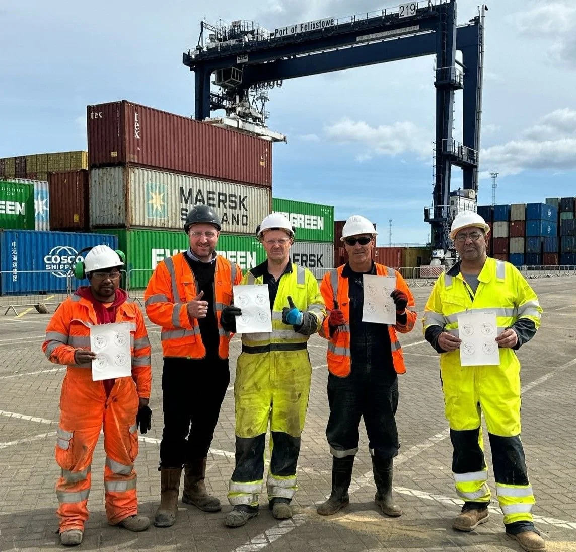 Group of five workers in safety gear holding papers with crane and shipping containers in background at Port of Felixstowe.