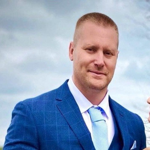 Man in a blue suit and light blue tie, standing outdoors with a cloudy sky background.