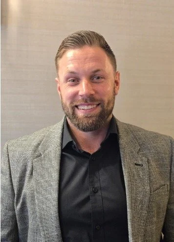 A smiling man with a beard and short hair, wearing a gray blazer and black shirt, standing in front of a neutral wall.