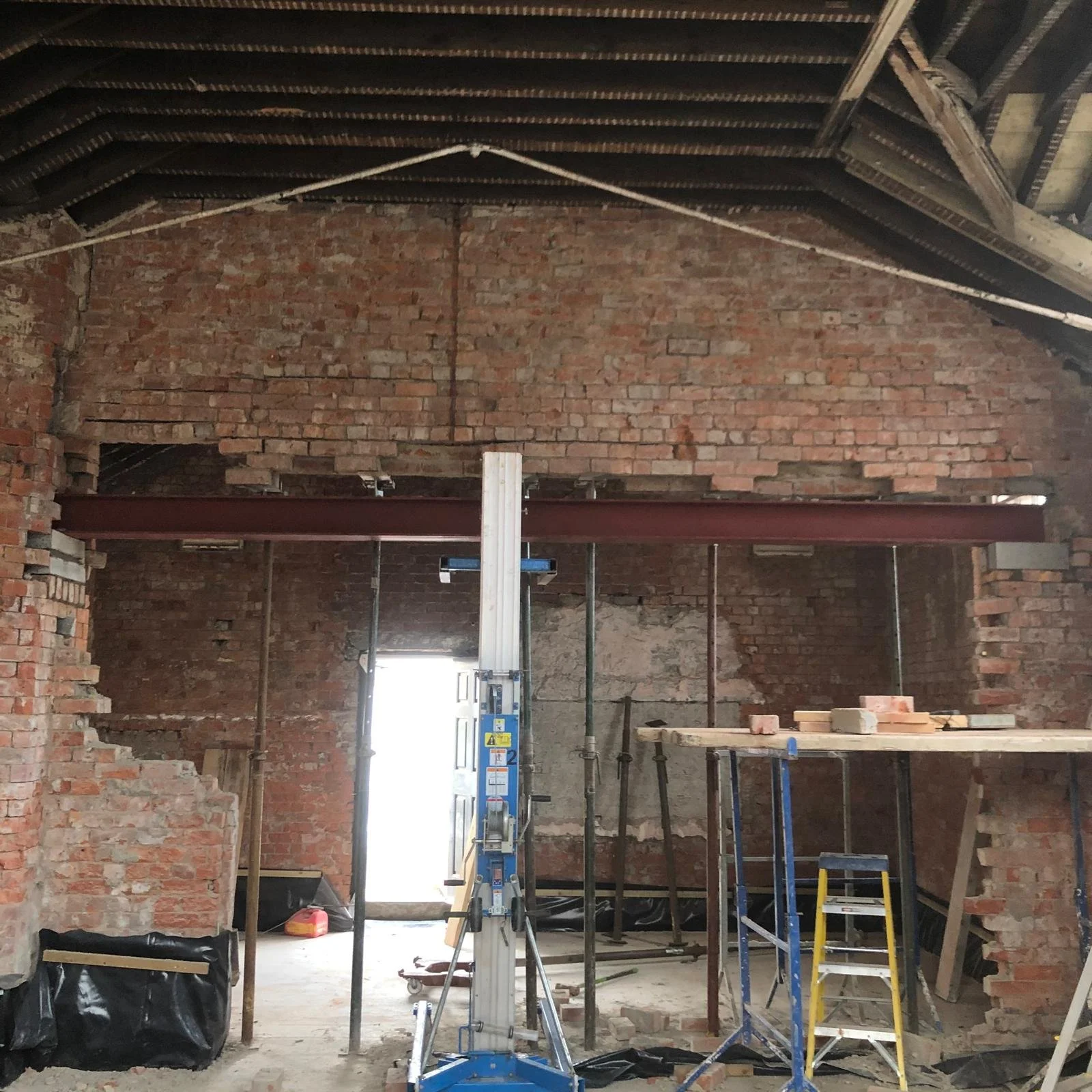 Interior of a partially renovated brick building, showing exposed brick walls, construction equipment, scaffolding, and a ladder.