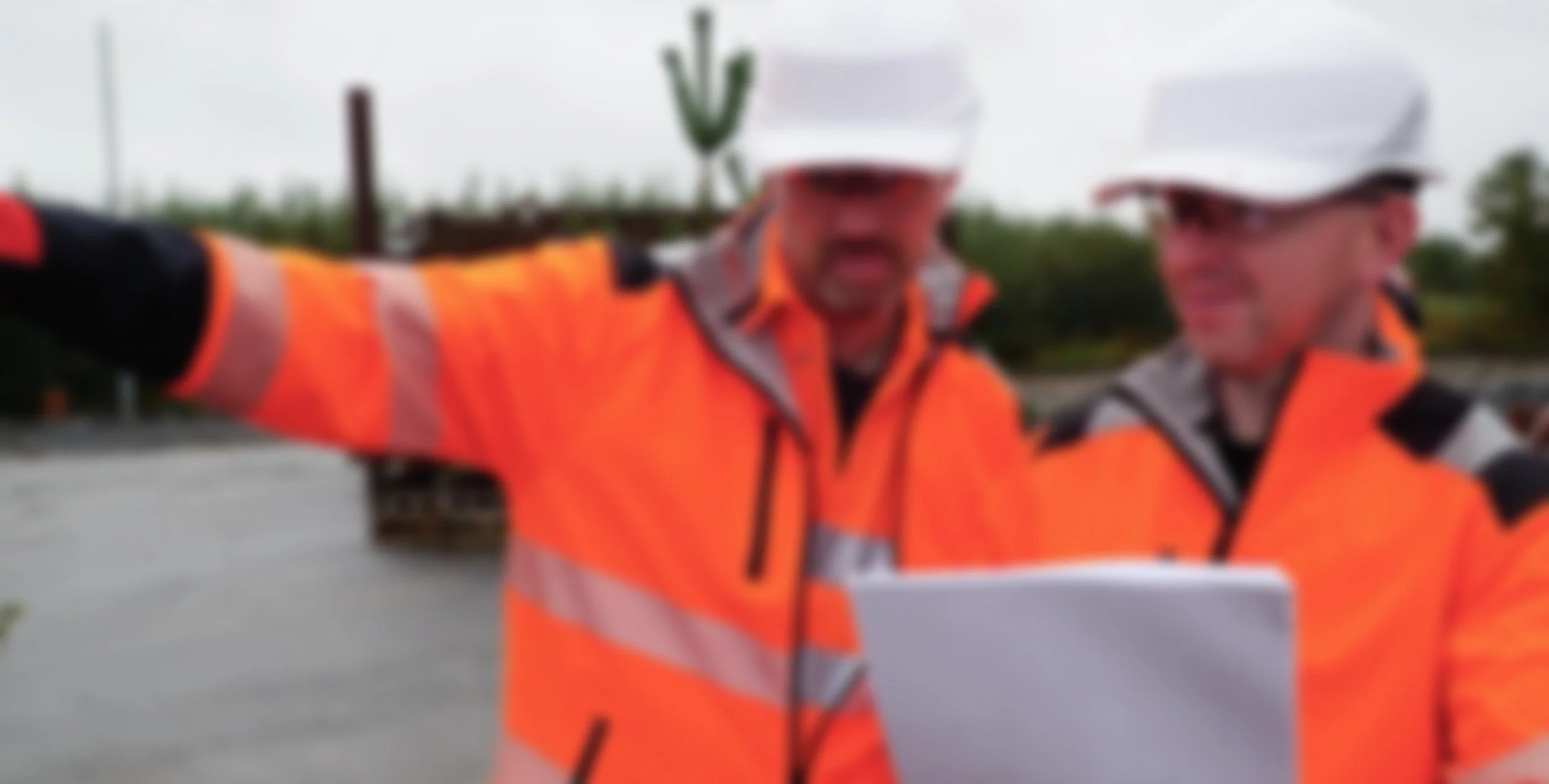 Two workers in orange safety vests and white hard hats reviewing a document outdoors.