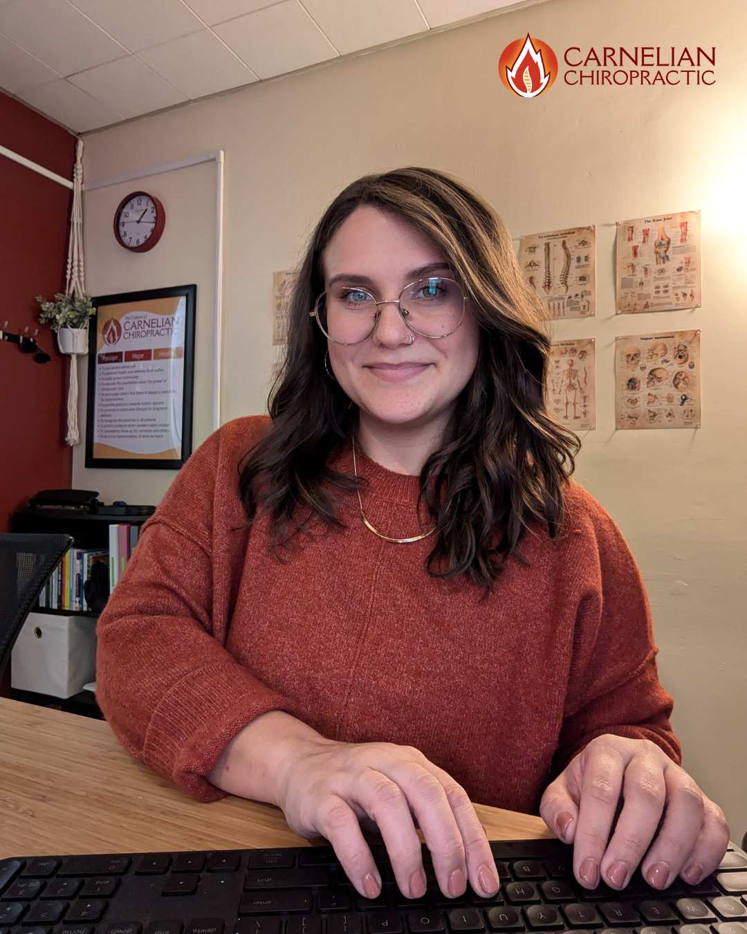 A woman with wavy brown hair and glasses, wearing a rust-colored sweater, sitting at a desk with a keyboard in front of her, inside a chiropractic office. The background shows medical charts on the wall, a clock, and a framed poster for Carnelian Chiropractic.