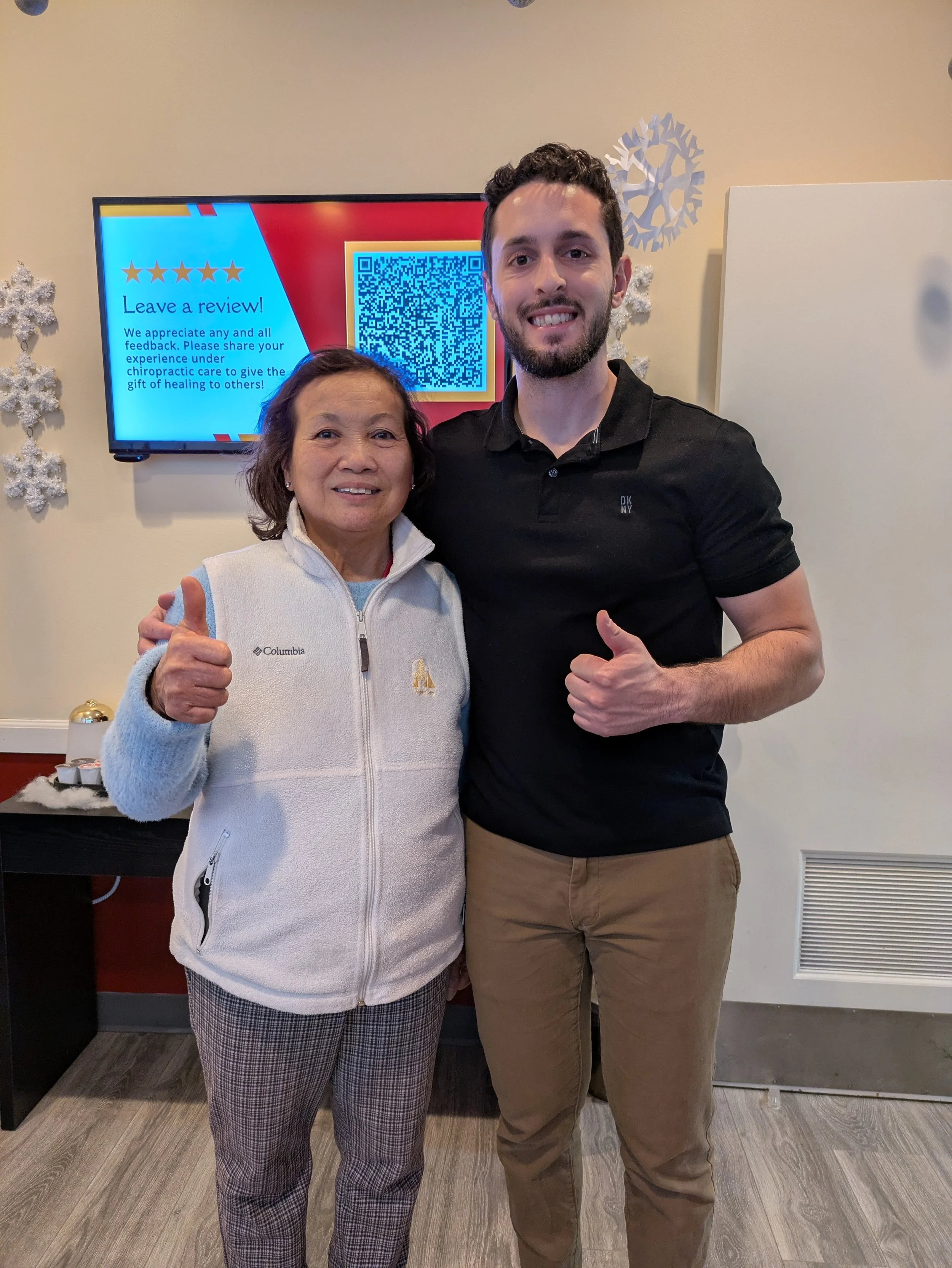 A smiling young man and an elderly woman giving thumbs up, standing indoors in front of a wall-mounted screen with a QR code and review prompt, decorated with snowflake wall hangings.
