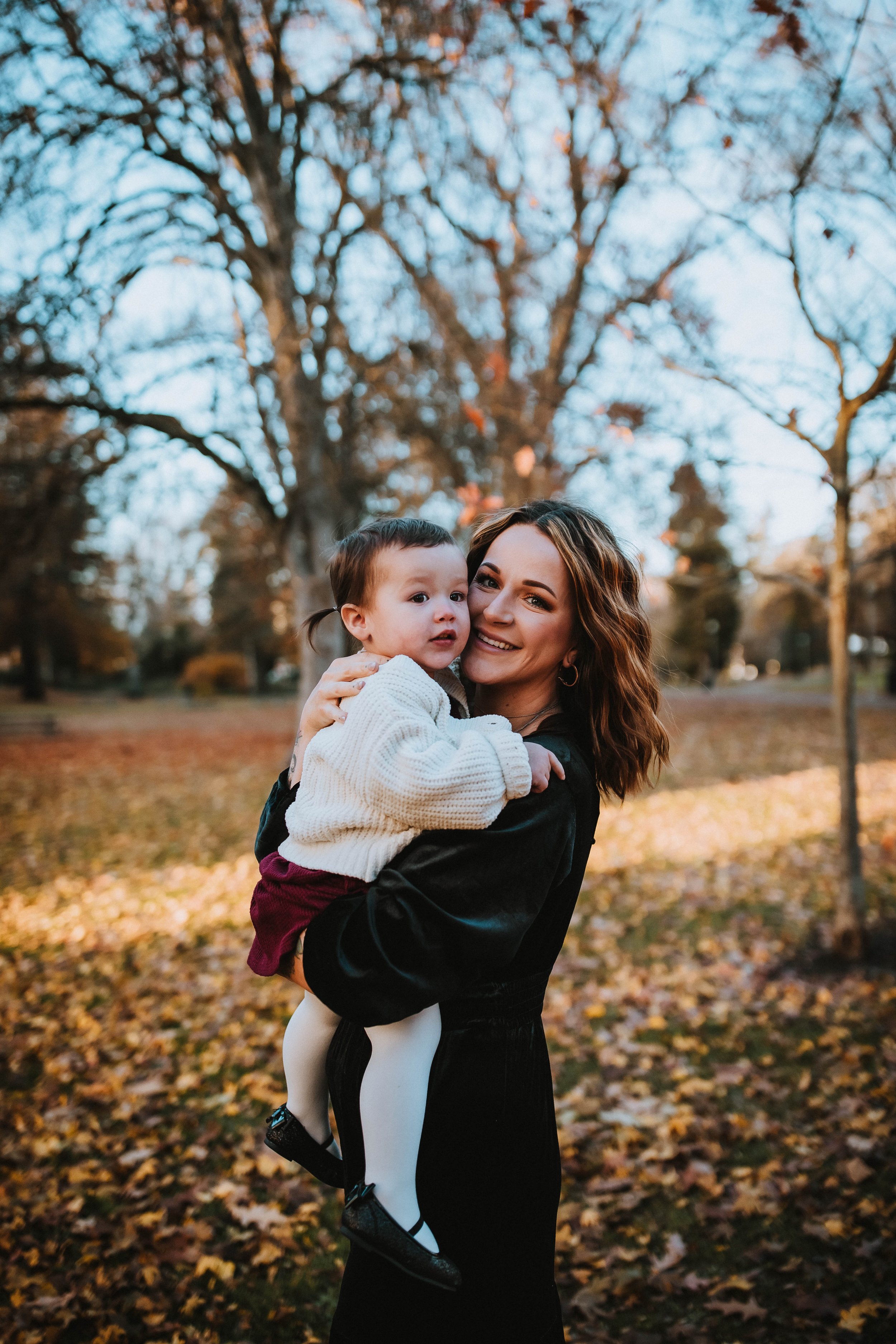 Woman holding a toddler in a park with autumn leaves on the ground.