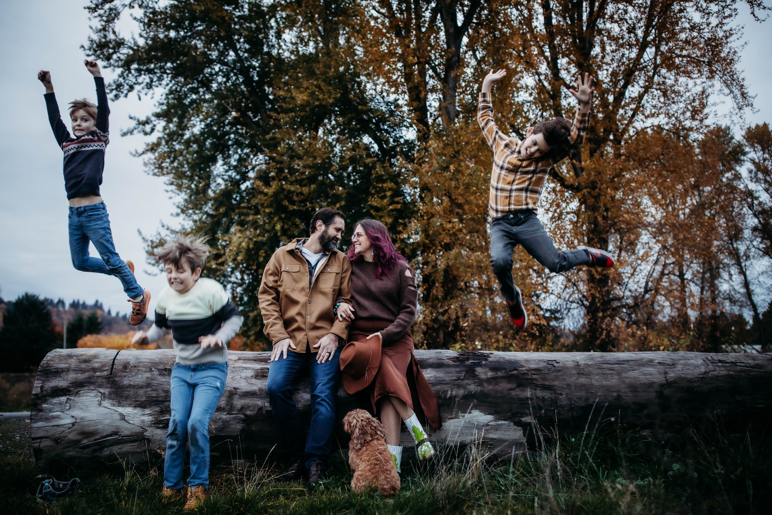 Family sitting on a log in a park, with children jumping and playing around, and a dog sitting nearby. Autumn trees in the background.