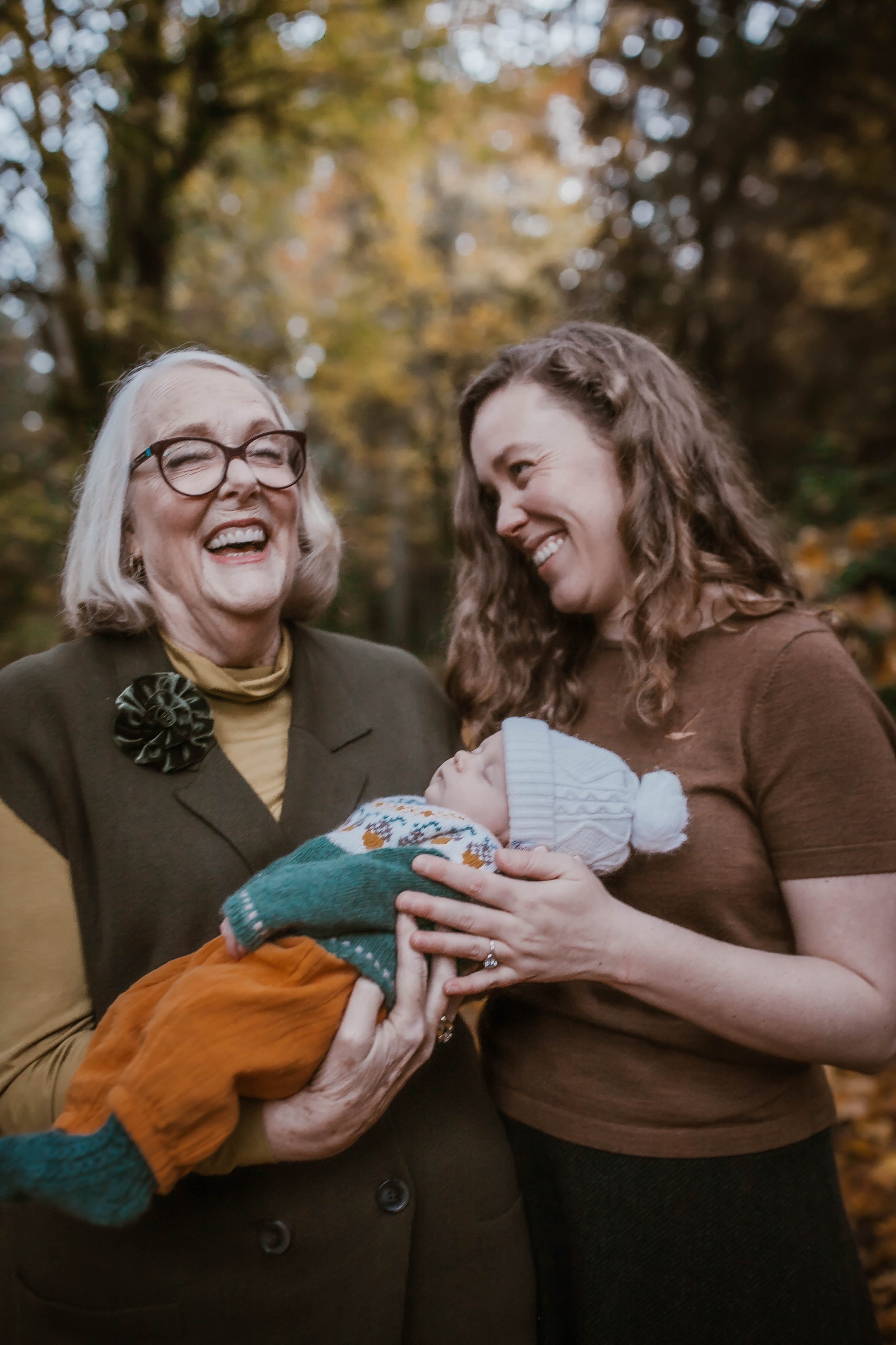 An older woman and a younger woman smiling while holding a baby wrapped in colorful clothing outdoors with a blurred forest background.