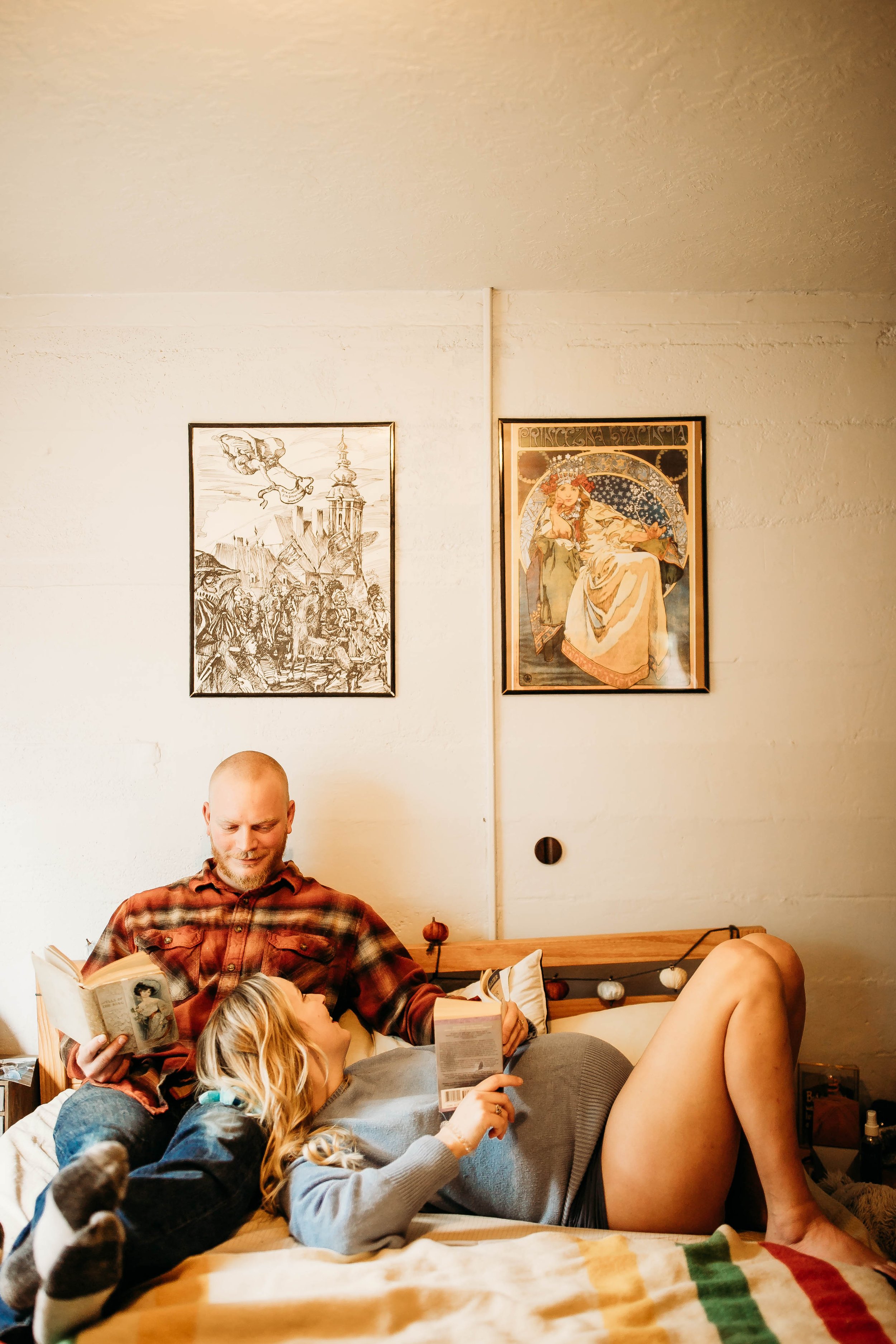 A couple relaxing on a bed in a cozy room. The man is wearing a plaid shirt and reading a book, while the woman, in a blue sweater, lays beside him with another book. Two framed artwork pieces are hung on the wall behind them. The bed has striped she