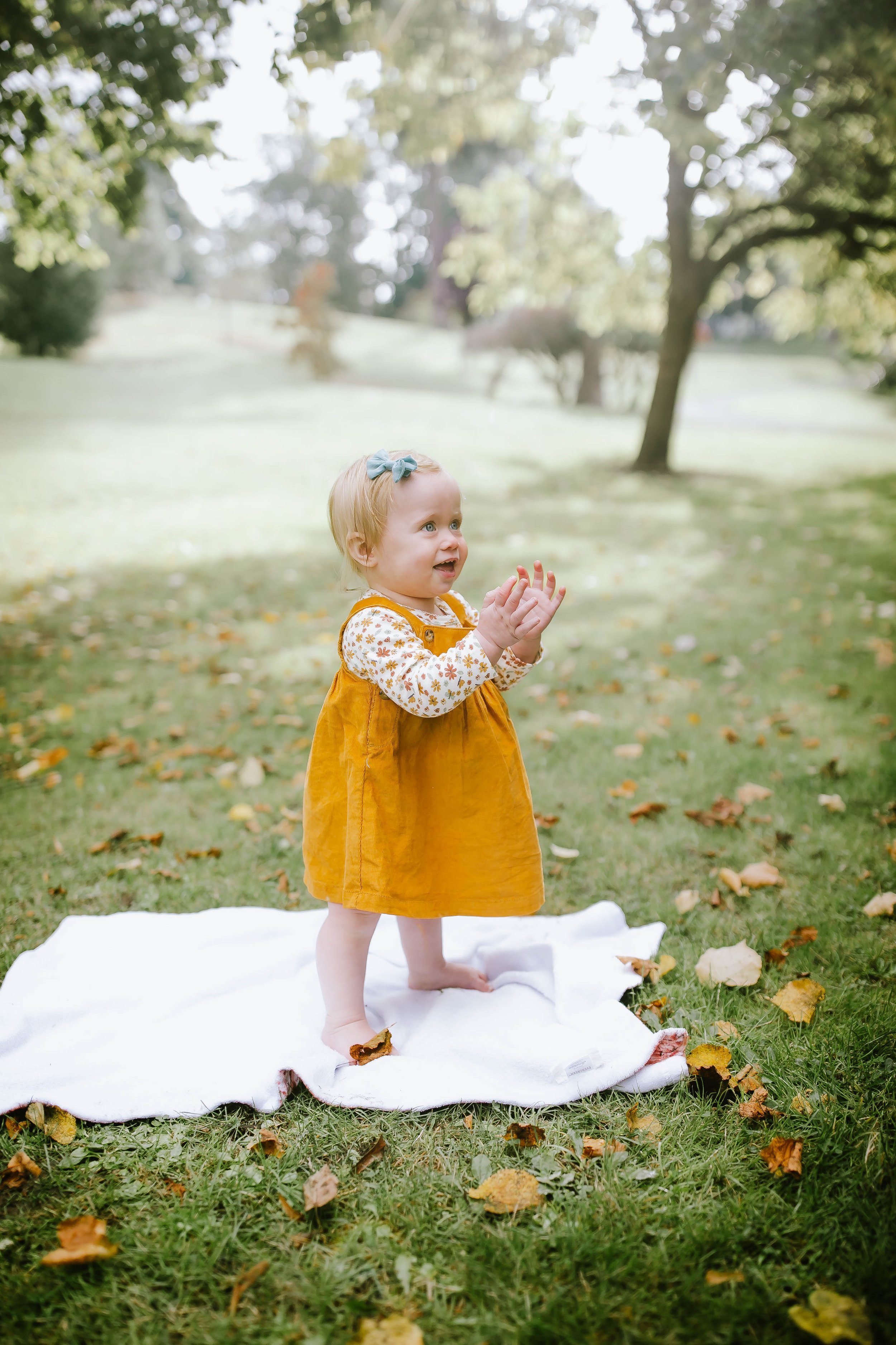 Young child in yellow dress standing on a white blanket outdoors with green grass and scattered leaves.