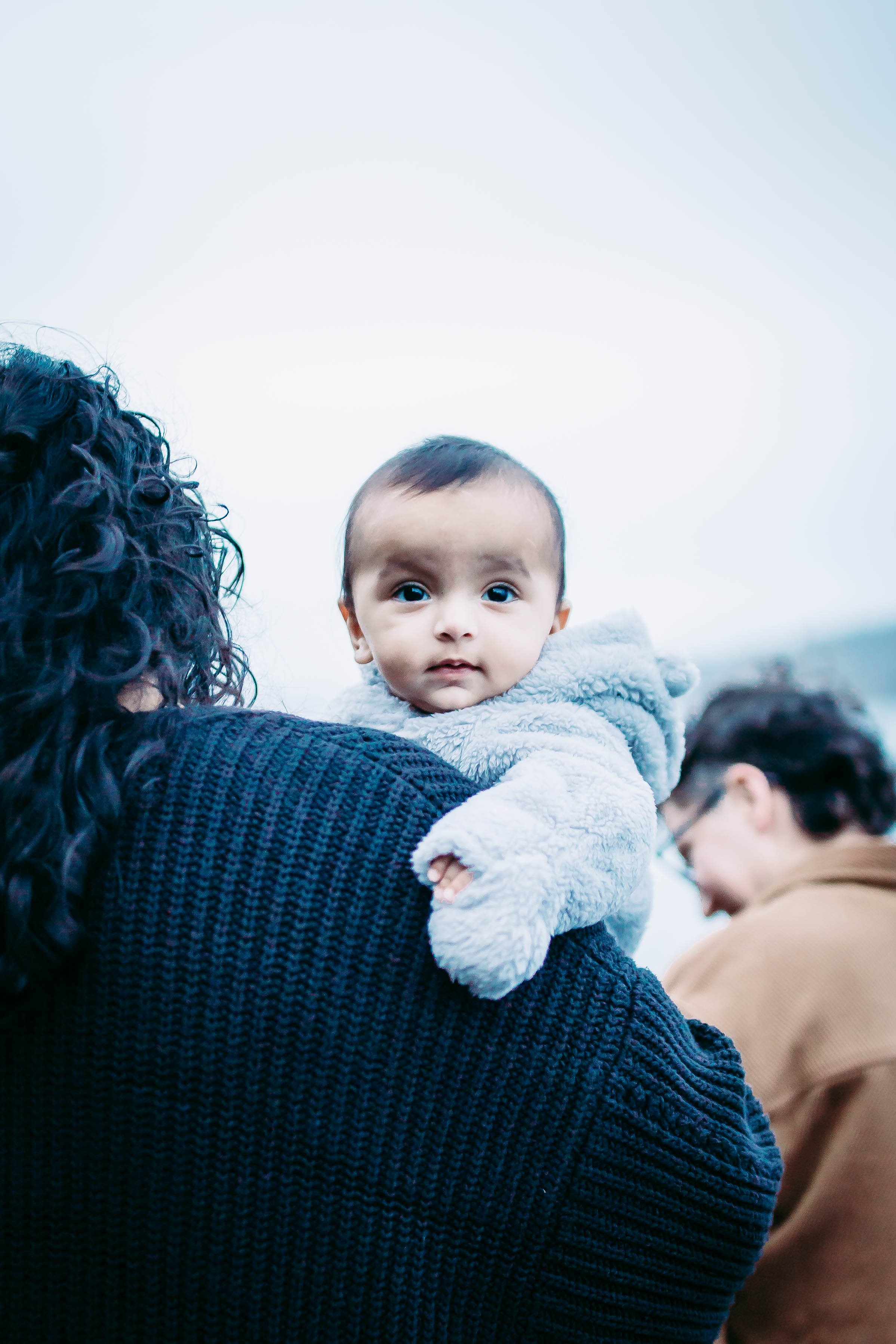 Baby in a gray outfit being held by a person wearing a dark sweater, with another person in a brown coat in the background.