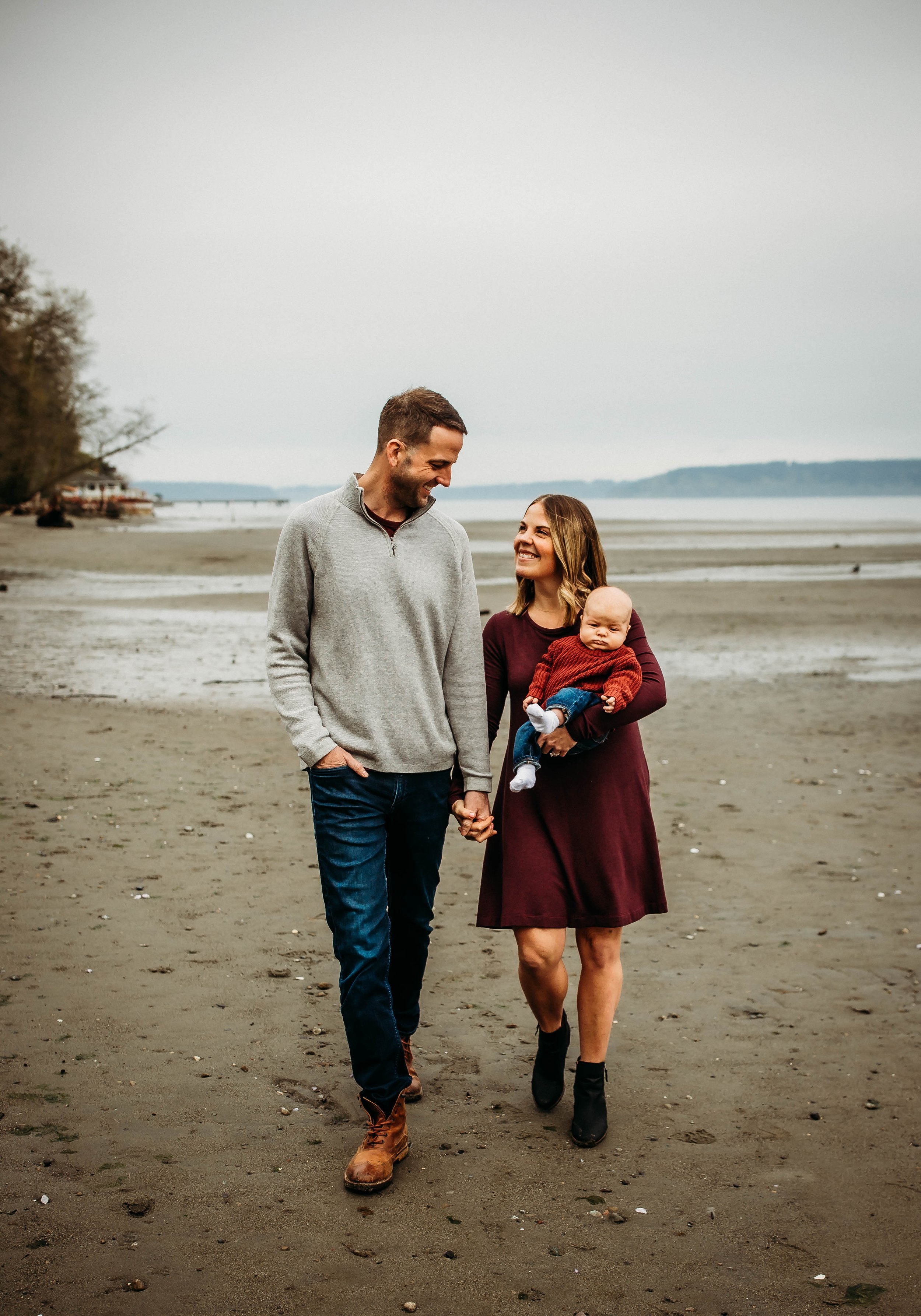 A man and woman walking on a beach, holding hands, with the woman carrying a baby. The sky is overcast and there are trees and water in the background.