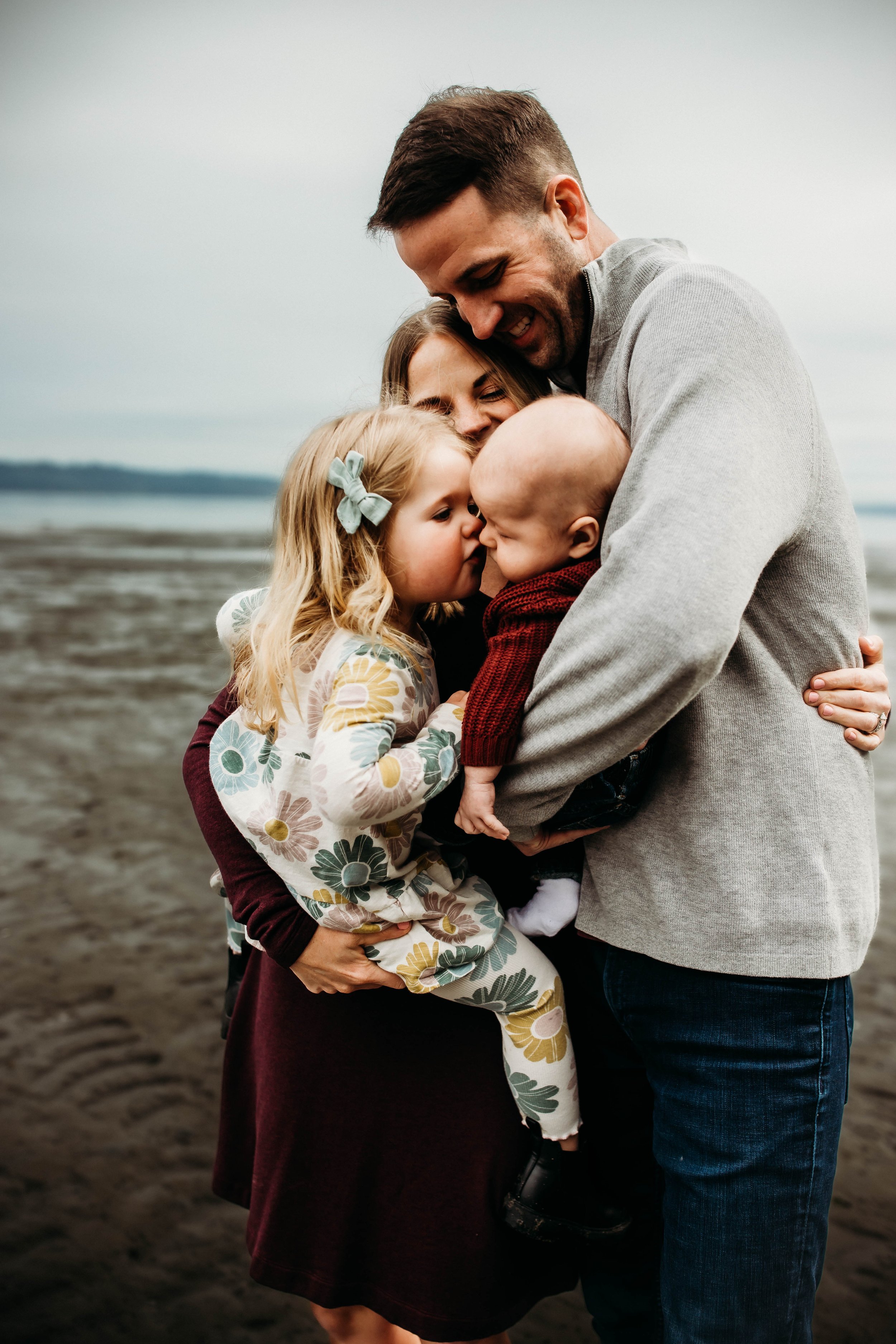 A family of four hugging on a beach, with a young girl kissing an infant held by the father, while the mother looks on smiling.