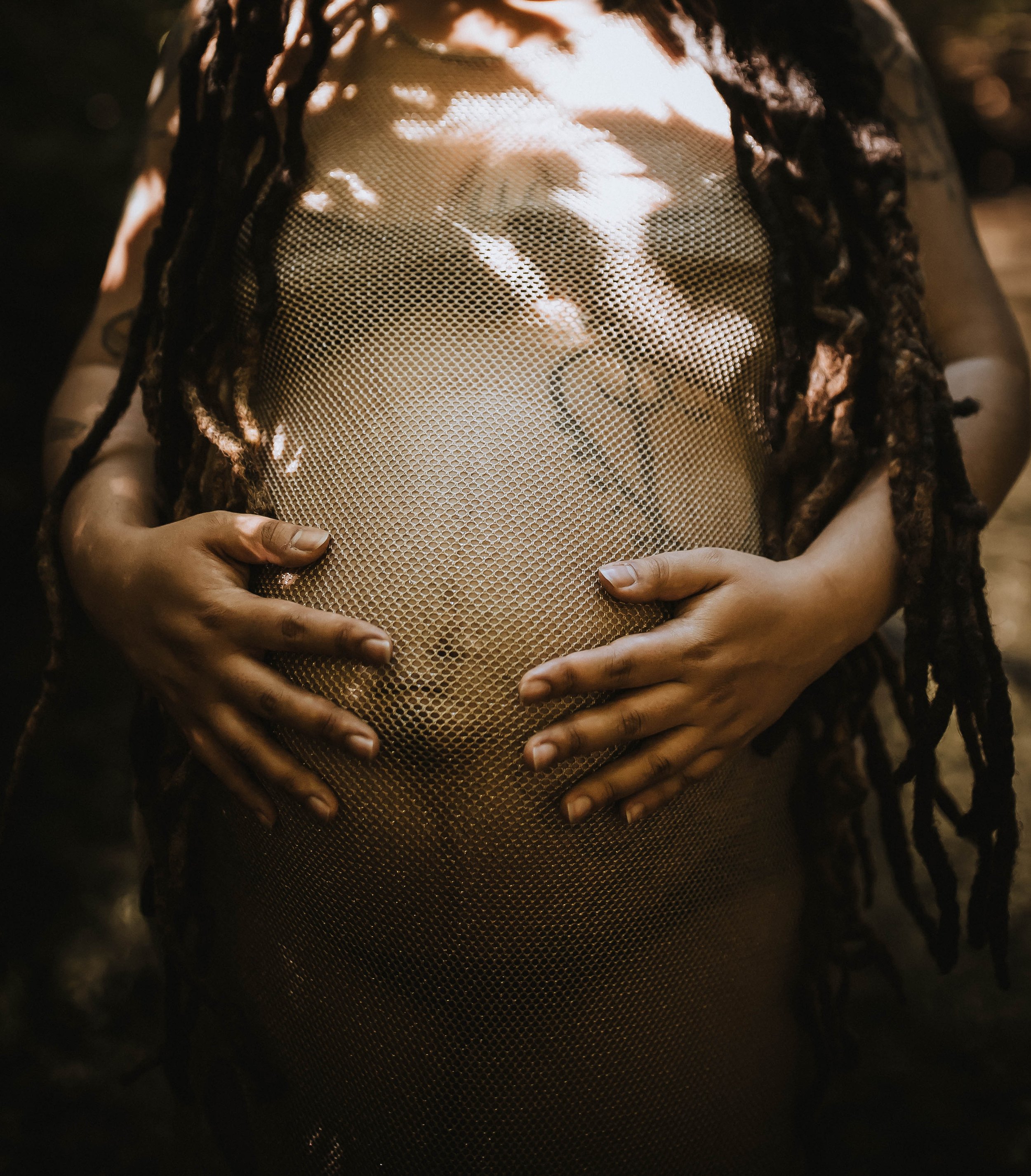 Pregnant person with dreadlocks touching their belly, wearing a mesh outfit, in a dappled sunlight setting.