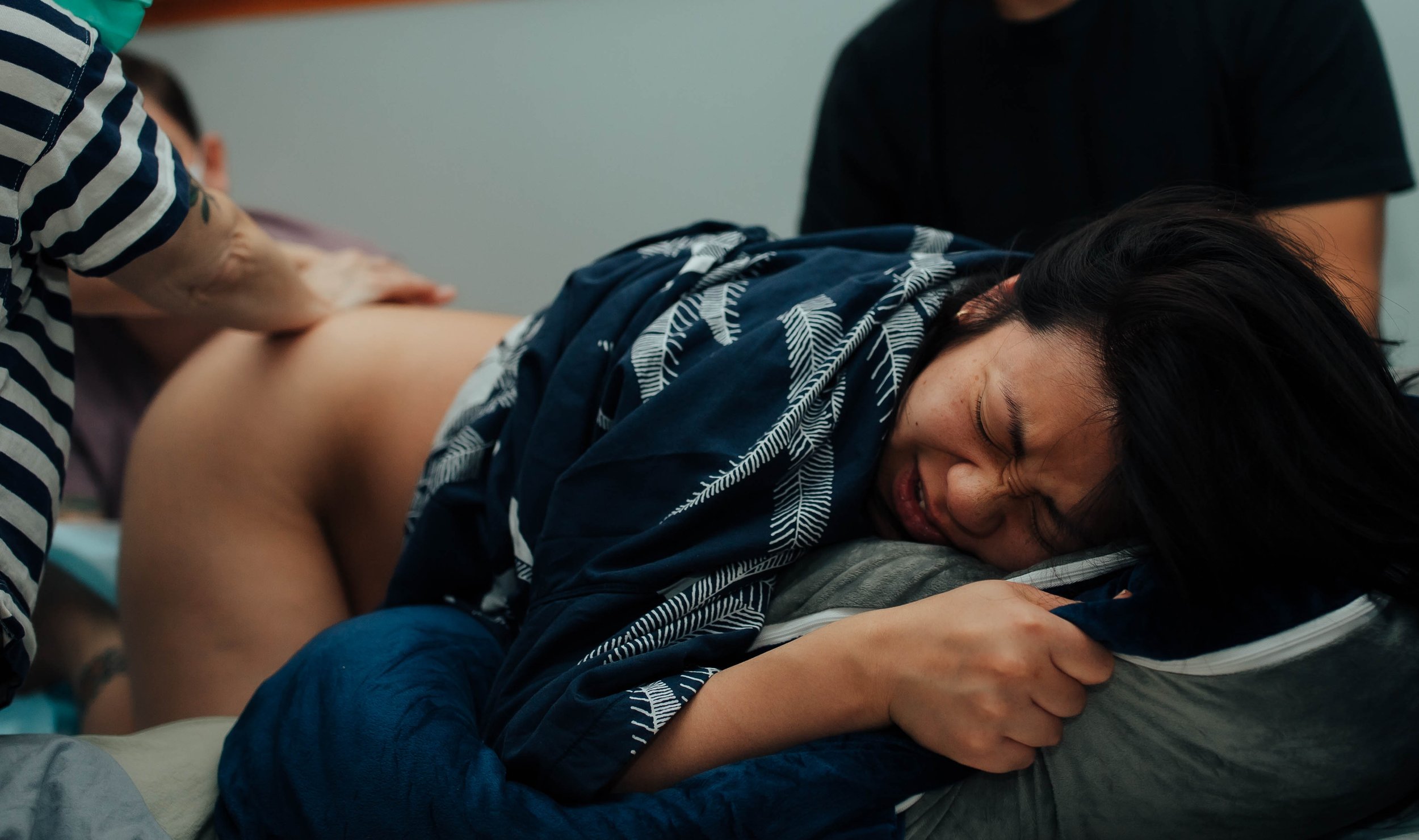 A person lying on a bed, appearing to be in labor, gritting their teeth and clutching a pillow. A caregiver is assisting by providing support, with a hand placed on the person's back.