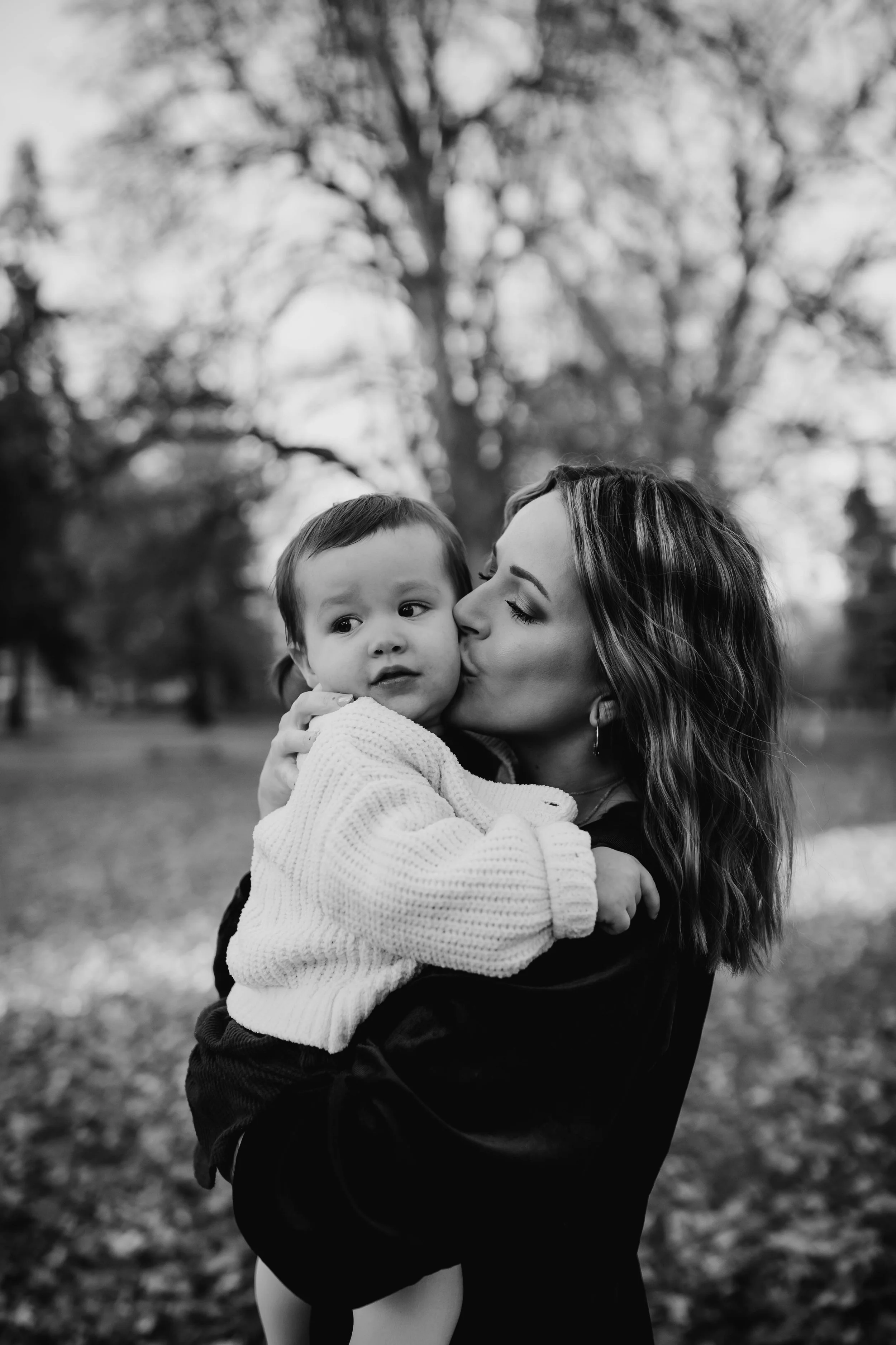 A black and white photo of a woman kissing a baby, holding the baby in her arms, with a blurred background of trees and foliage.