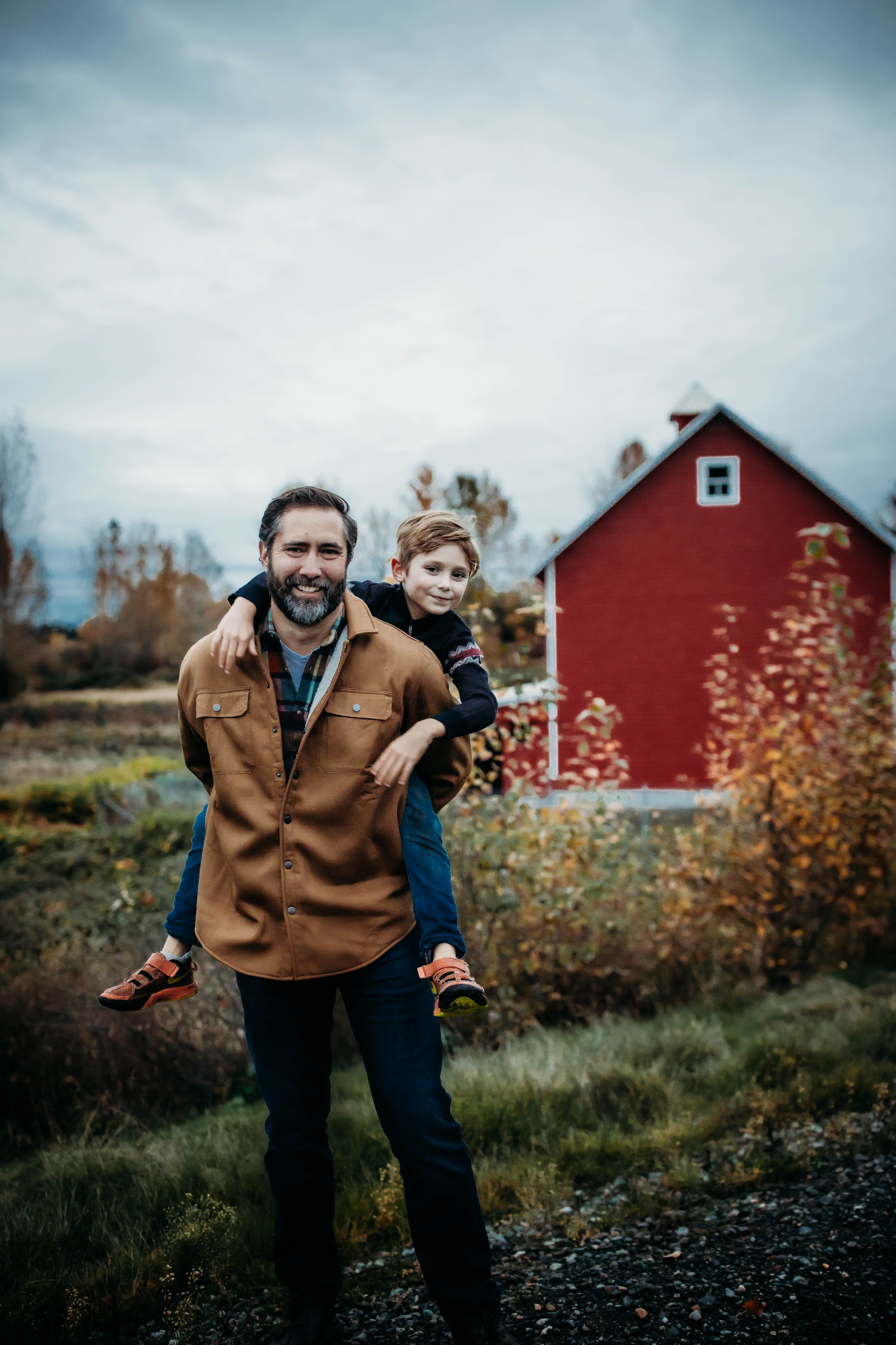 Man carrying a child on his back in front of a red barn, with autumn foliage in the background.