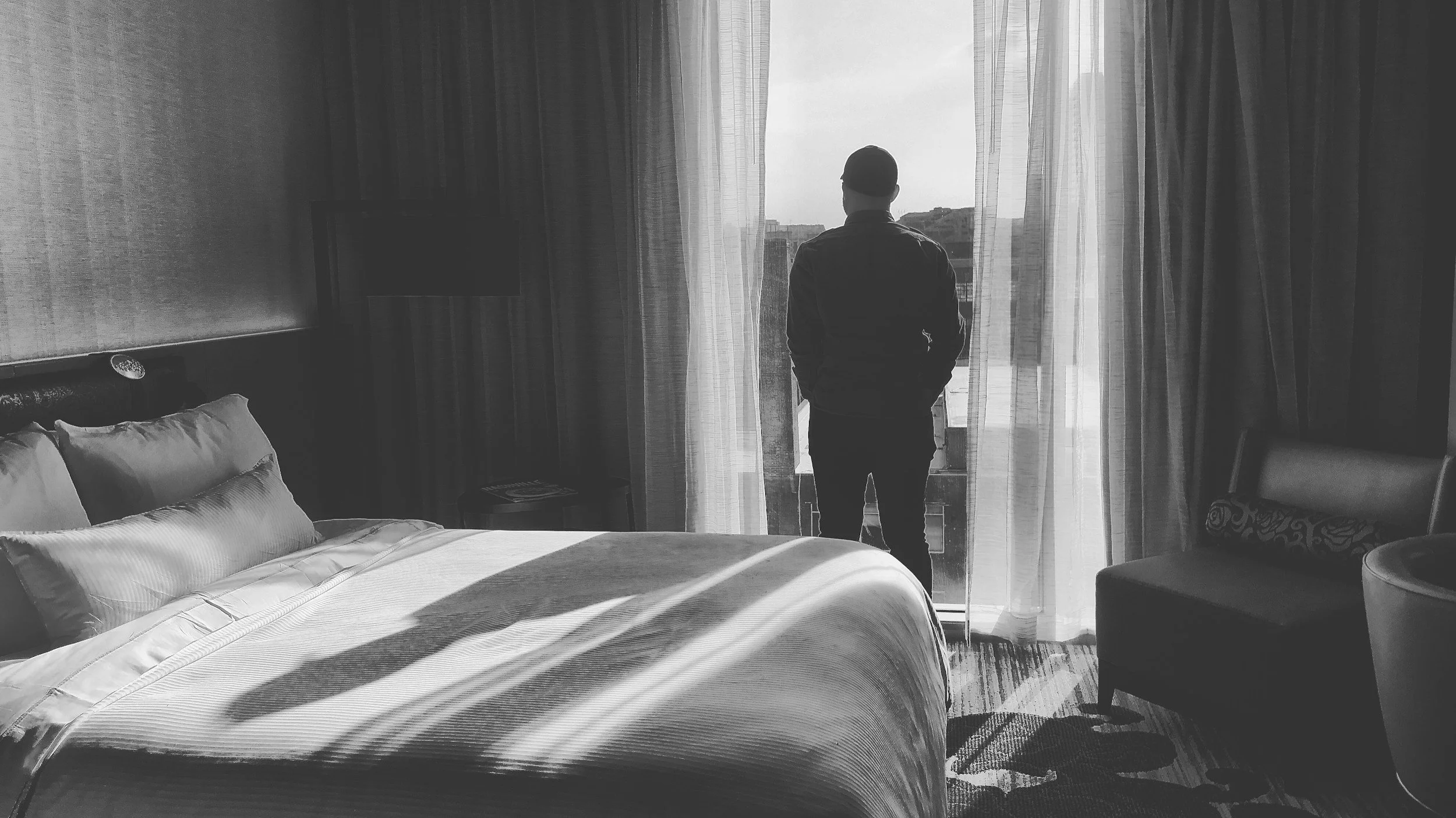 A person with short hair, wearing a jacket and cap, standing in a hotel room near a large window with curtains drawn open, looking outside. The room has a bed with pillows and a chair, with sunlight casting shadows across the bed.