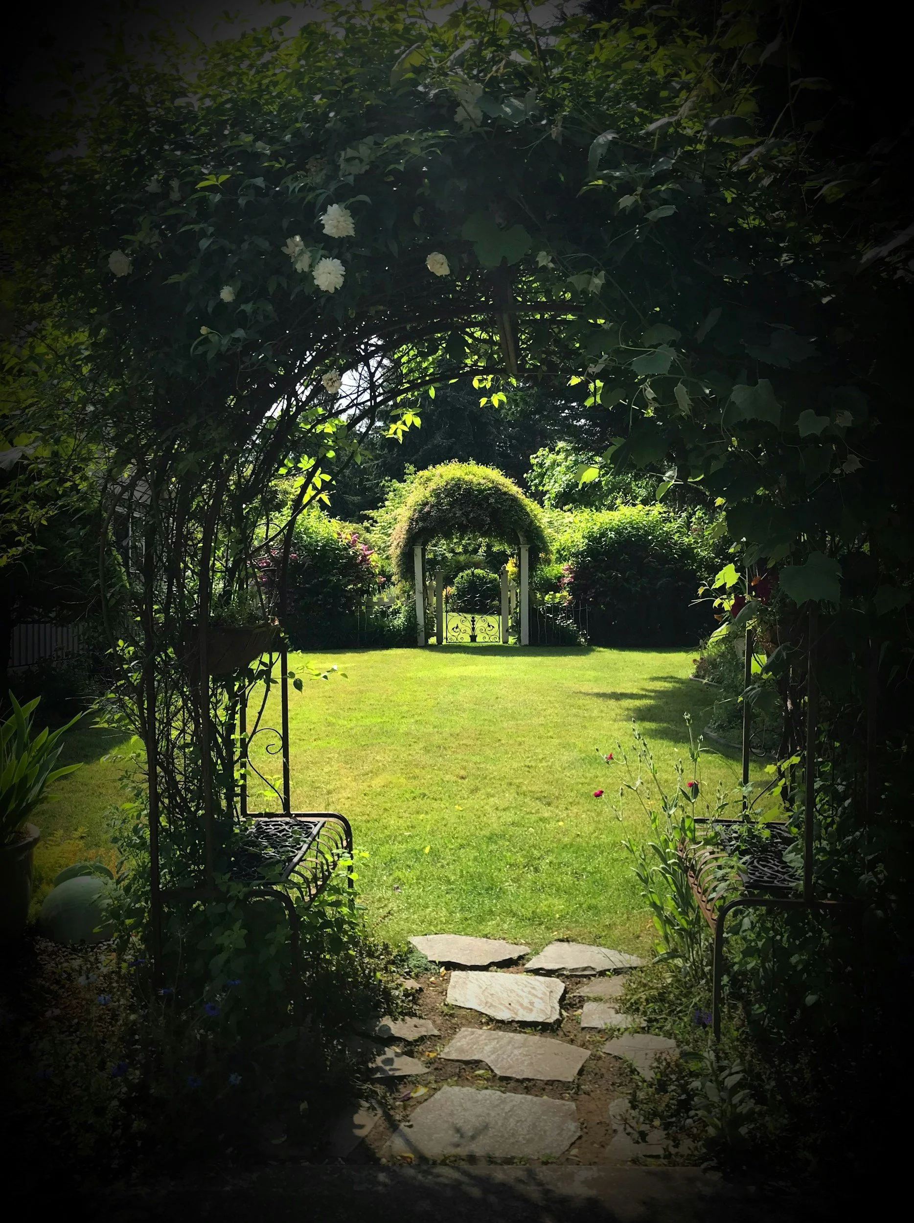 View through a garden archway leading to a lush, green lawn with a decorative gated arbor in the background, surrounded by flowering bushes and dense foliage.