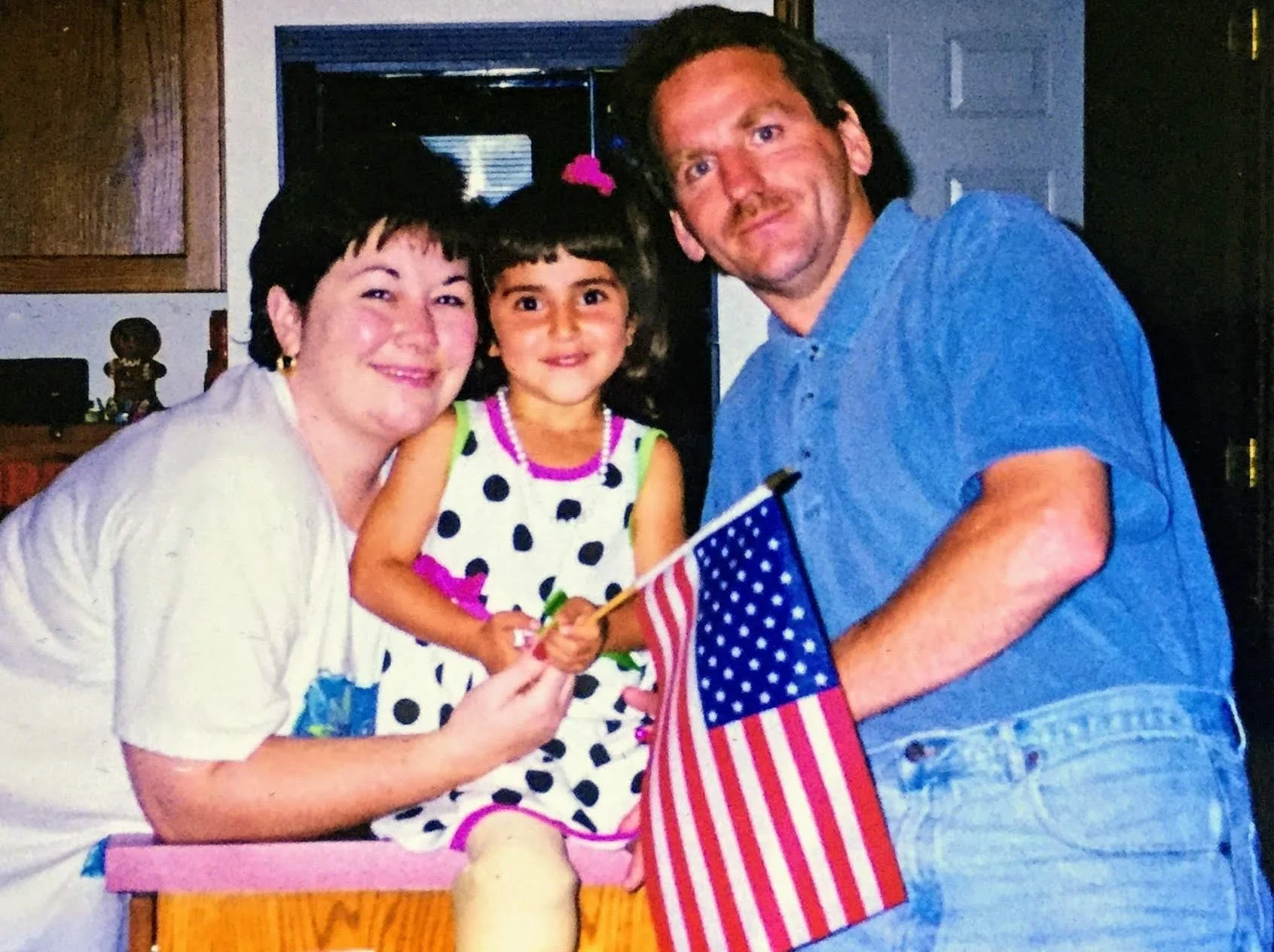 Toddler holding an American Flag with her new adoptive parents on either side of her.